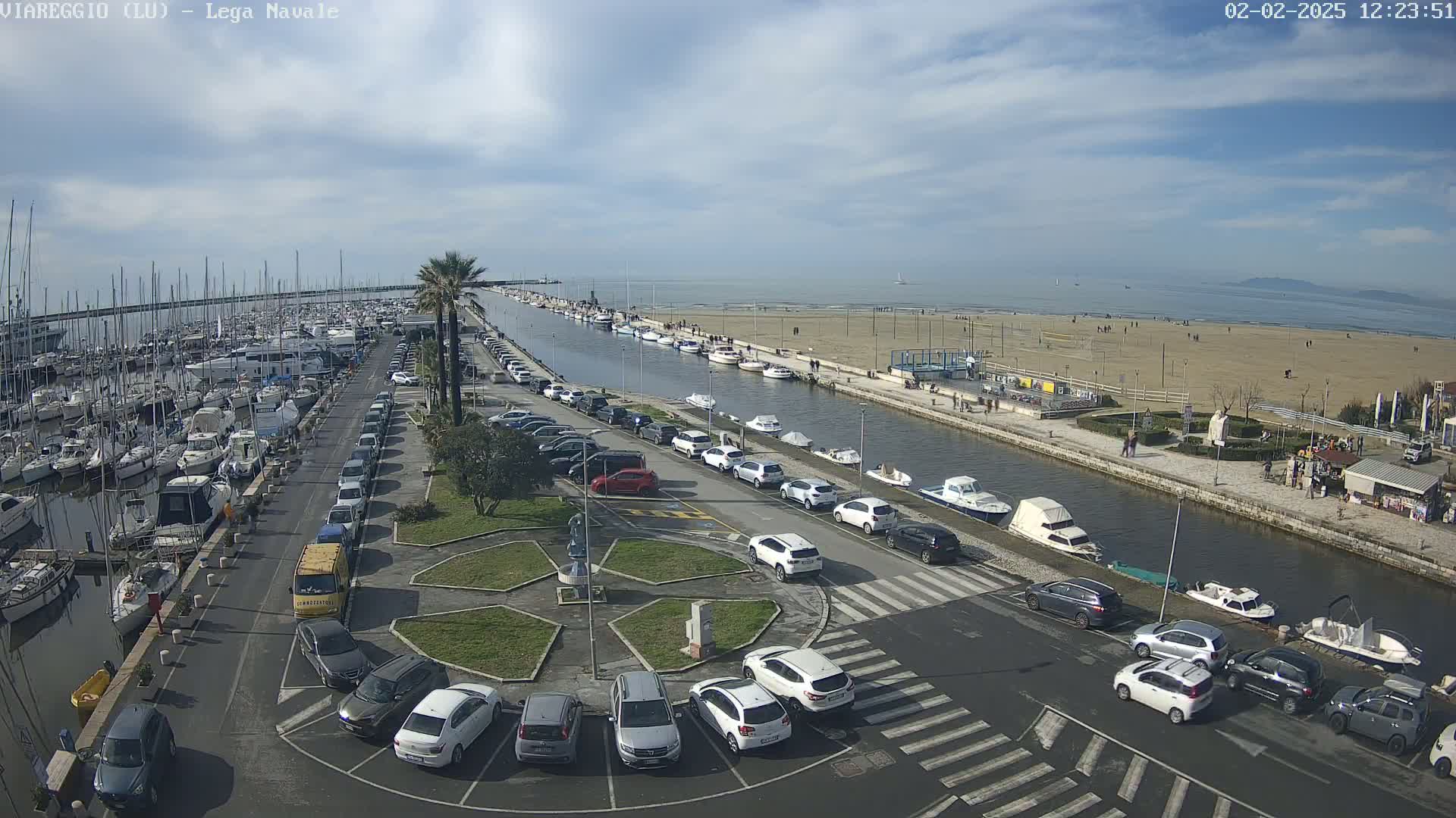 A marina filled with boats and a parking lot next to a canal leading to a sandy beach under a partly cloudy sky.