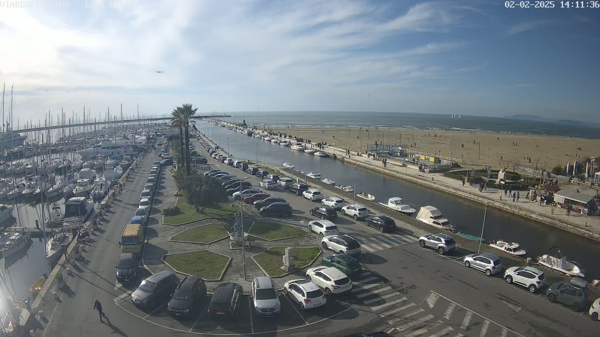 A marina filled with boats is adjacent to a canal and a sandy beach on a sunny day.