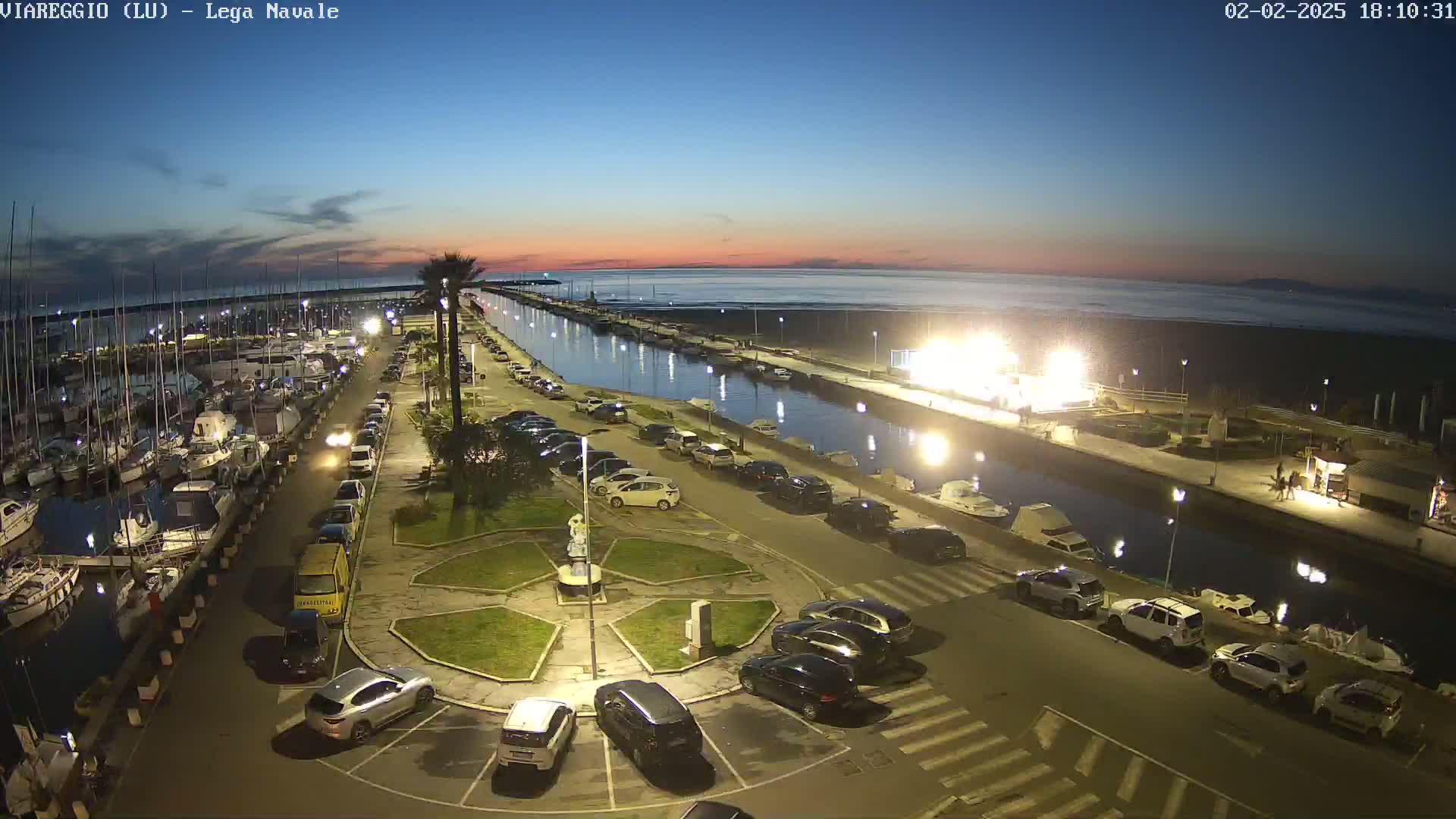 An aerial nighttime view of a marina with numerous boats, cars parked along a canal, and a beach in the background under a twilight sky.