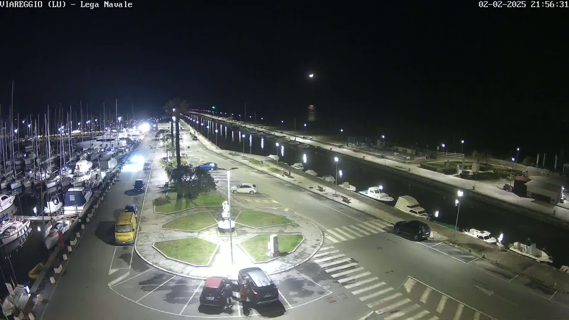 A nighttime view of a marina with many boats docked, a roadway with parked cars, and a small roundabout in the center, under a mostly clear sky with a visible moon.