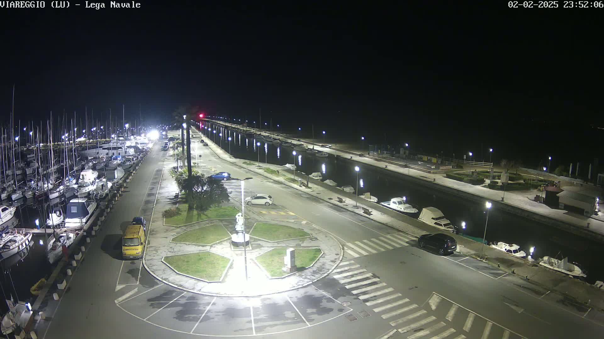 A marina at night, illuminated by streetlights, shows numerous boats docked along a waterfront, with cars parked in a nearby lot and a small roundabout featuring a statue in the center.