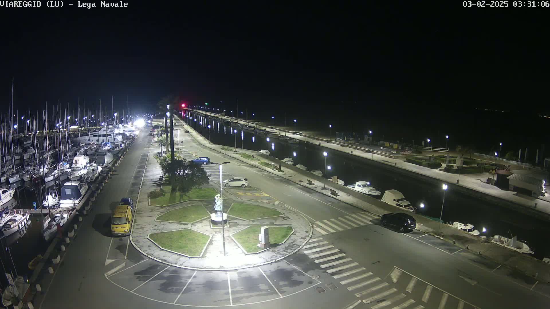 A nighttime, aerial view of a marina with numerous boats docked, a roadway with parked cars, and a circular plaza featuring a statue, all under clear skies.