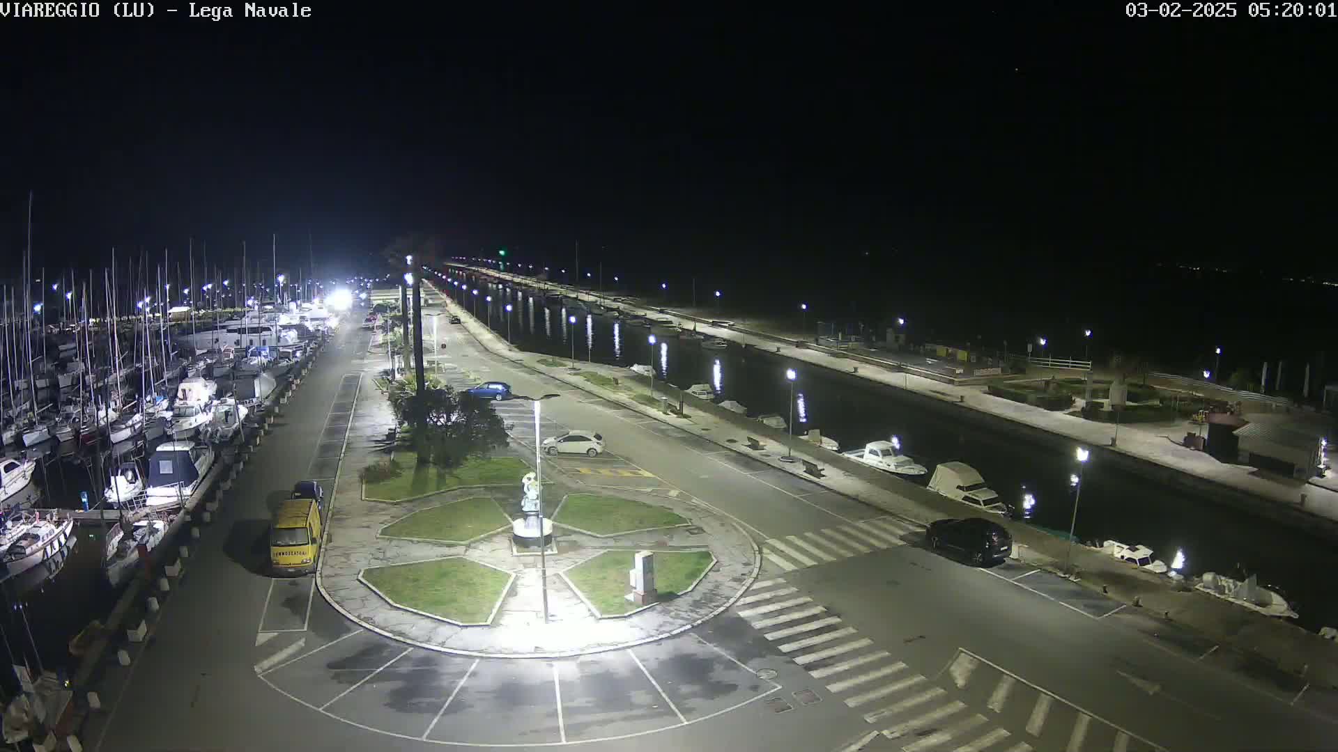 A nighttime aerial view of a marina with numerous boats docked, a roadway with parked cars, and a central roundabout featuring a statue, all brightly lit under clear skies.
