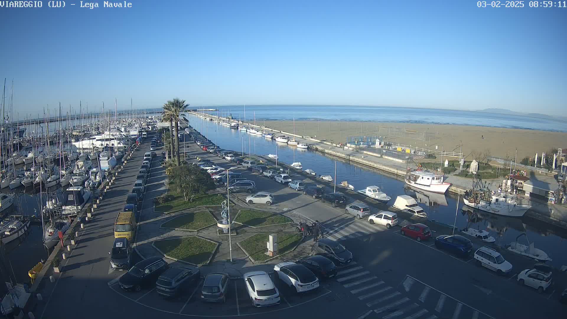 A marina filled with sailboats and motorboats is next to a canal and a sandy beach under a clear, sunny sky.