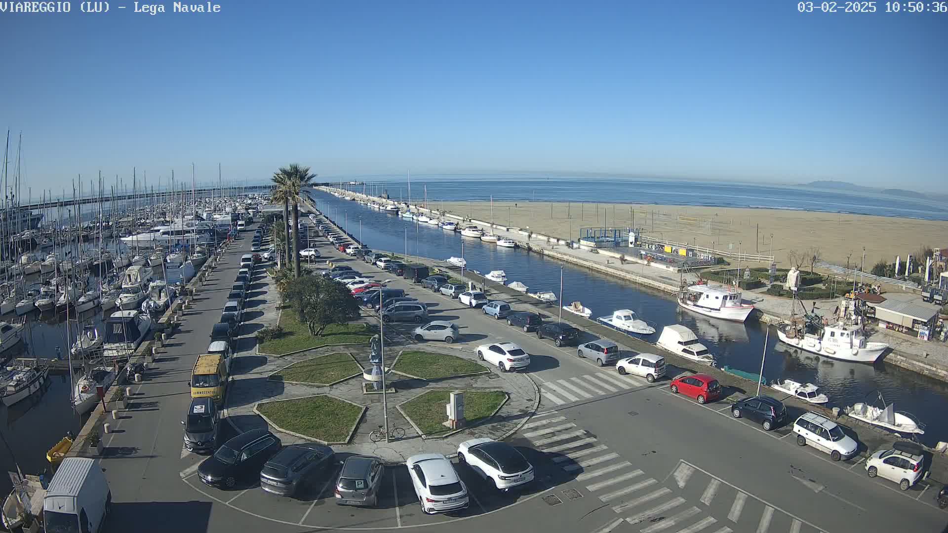 A marina filled with boats is next to a canal with parked cars and a sandy beach under a clear blue sky.