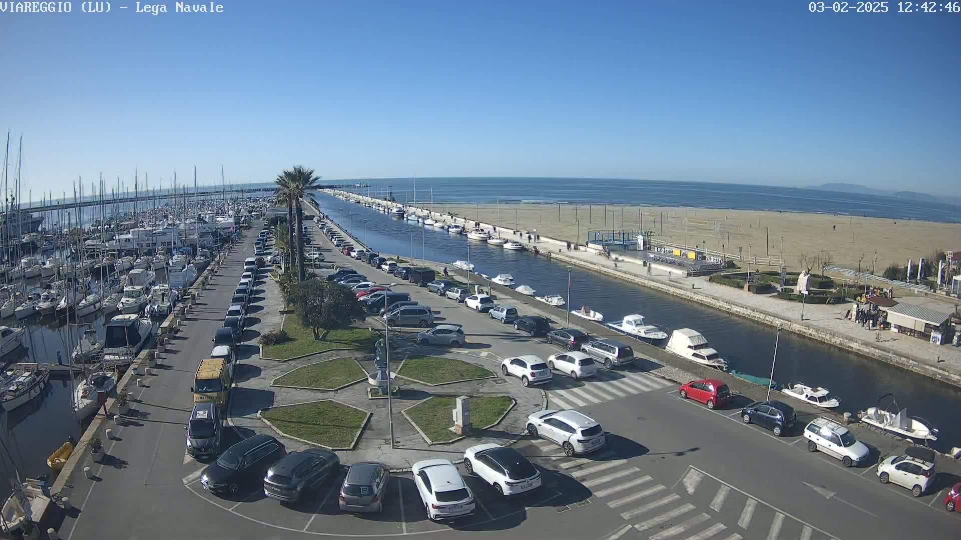 A marina filled with boats is adjacent to a parking lot and canal, which leads to a sandy beach under a clear blue sky.