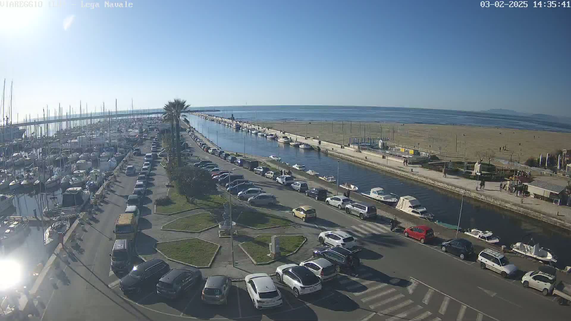 A harbor filled with boats is adjacent to a parking lot and canal, all overlooking a sandy beach and calm ocean under a clear, sunny sky.