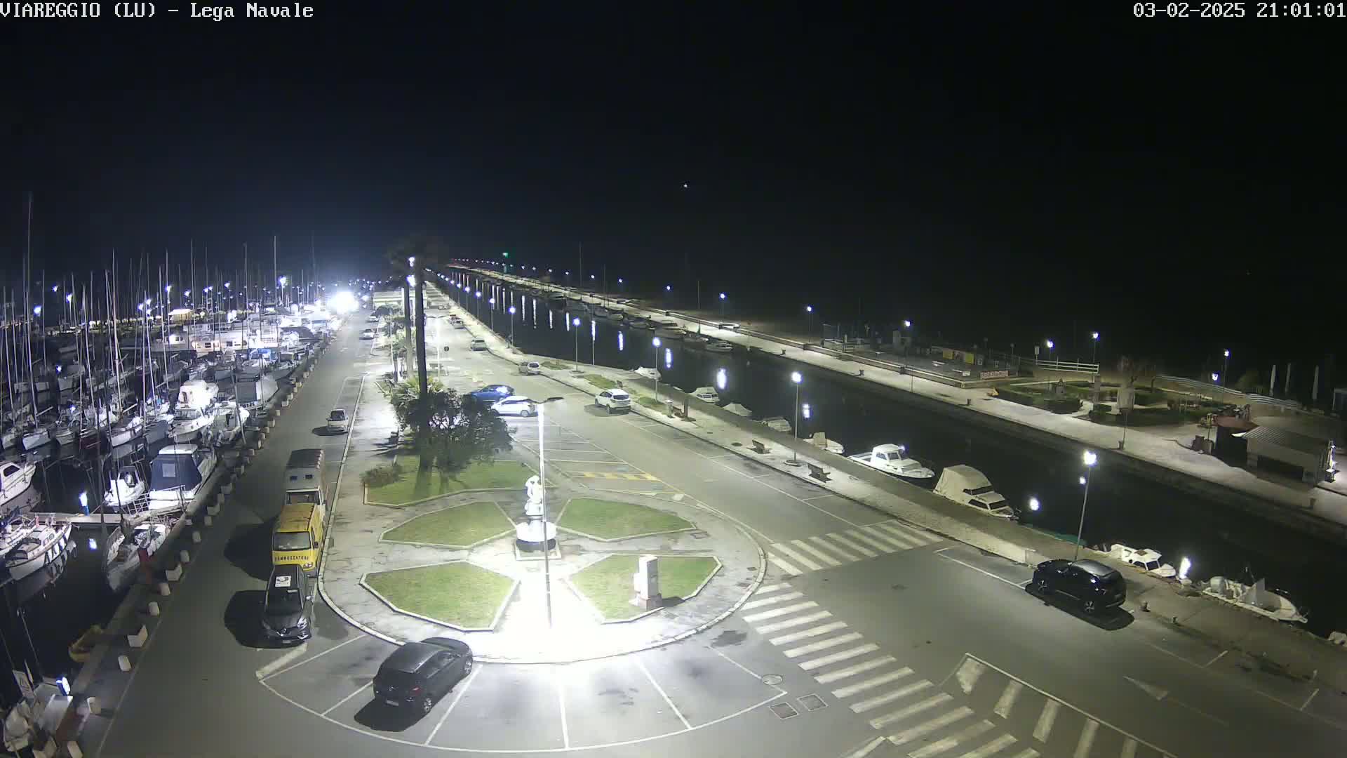 A nighttime aerial view of a marina with numerous boats docked, a roadway lined with parked cars, and a central roundabout with landscaping.