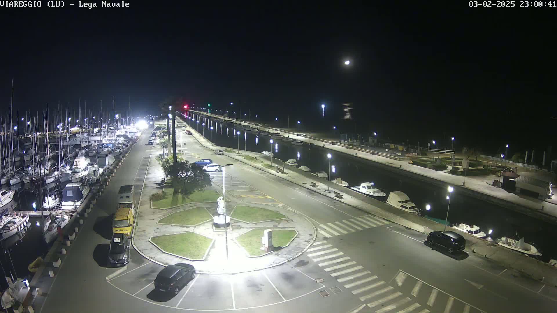 A nighttime aerial view of a marina with numerous boats docked, a roadway with parked cars, and a central roundabout, under a clear moonlit sky.