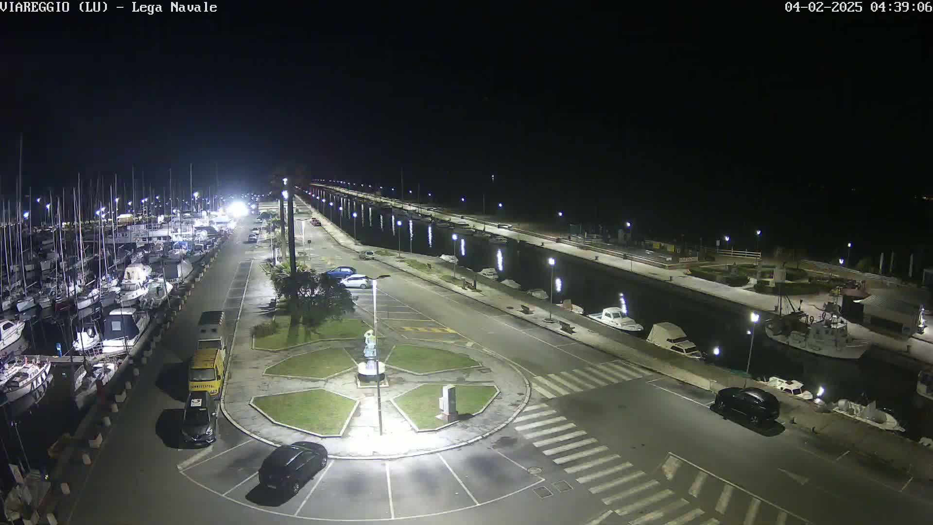 A nighttime, aerial view of a marina with numerous boats docked, a roadway with cars parked, and a small, central plaza featuring a statue.