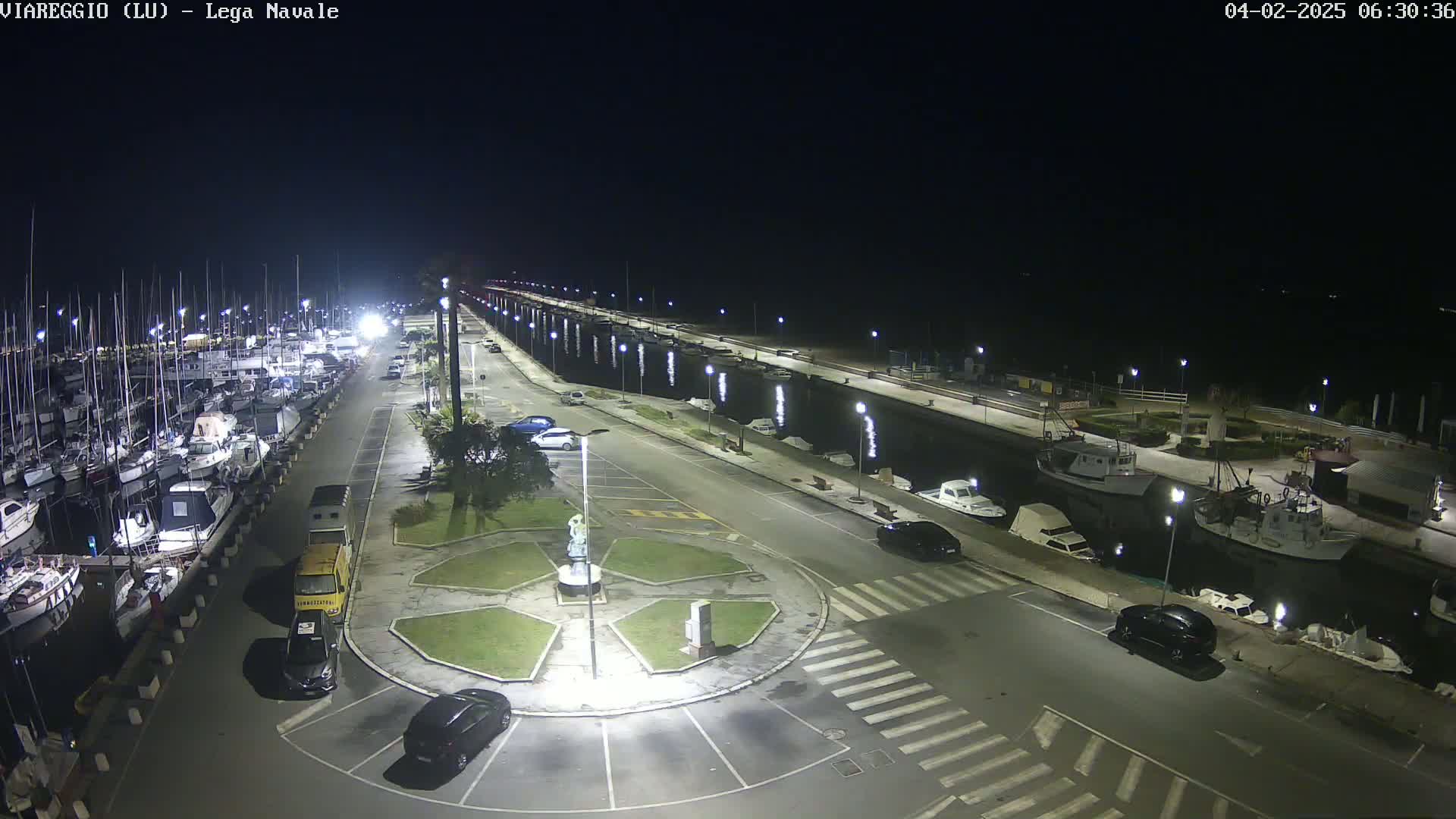 A nighttime aerial view of a marina with numerous boats docked, a roadway with parked cars, and a small plaza featuring a statue in the center.