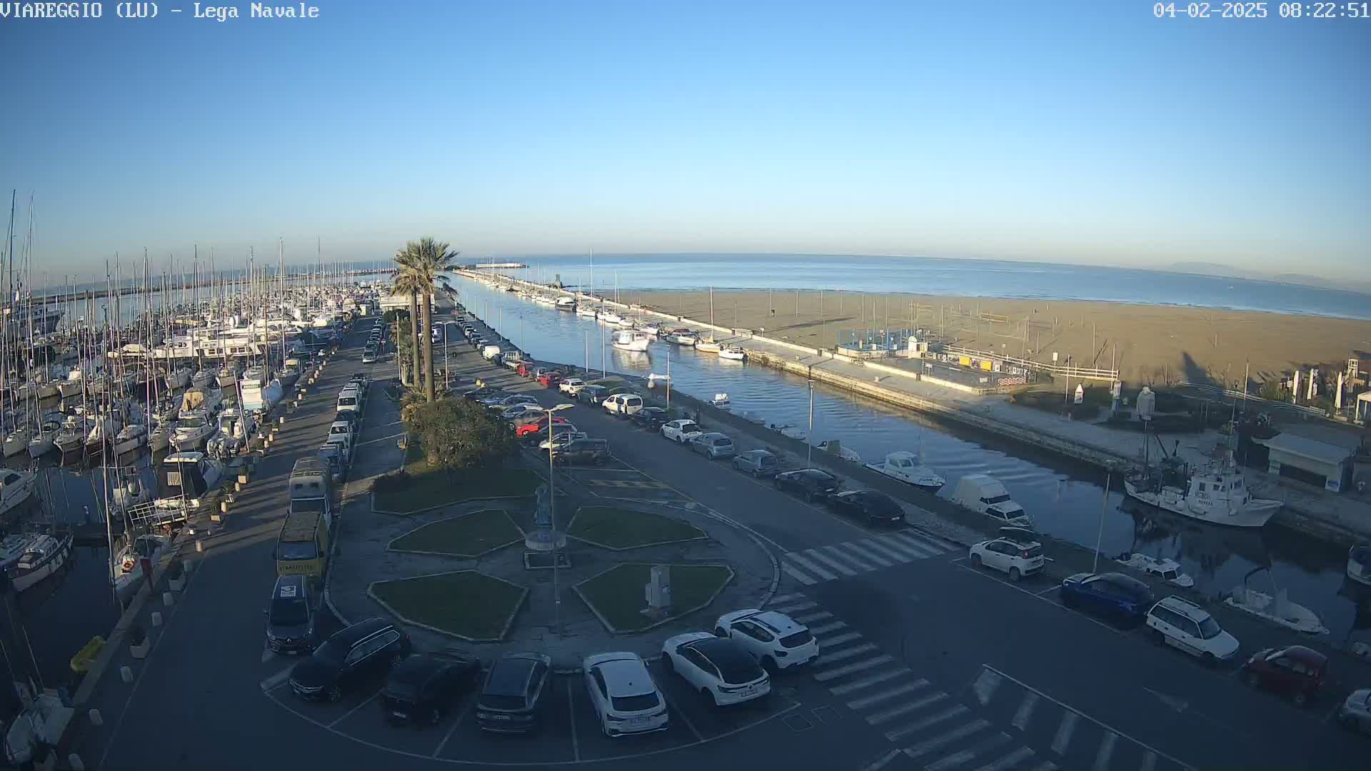 A marina filled with boats is adjacent to a sandy beach and calm ocean under a clear blue sky.