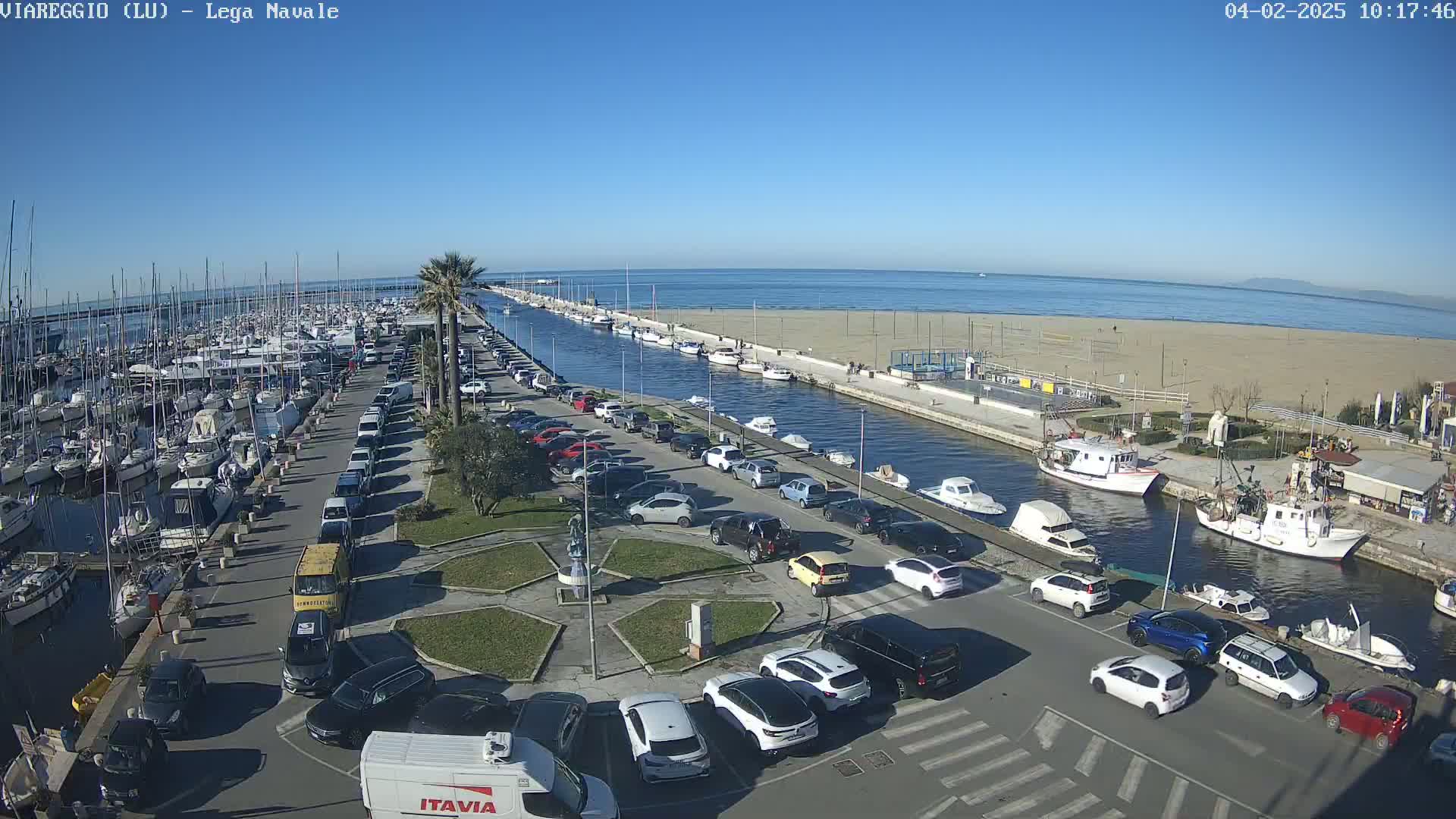 A marina filled with boats is adjacent to a sandy beach and canal with cars parked alongside, all under a clear blue sky.