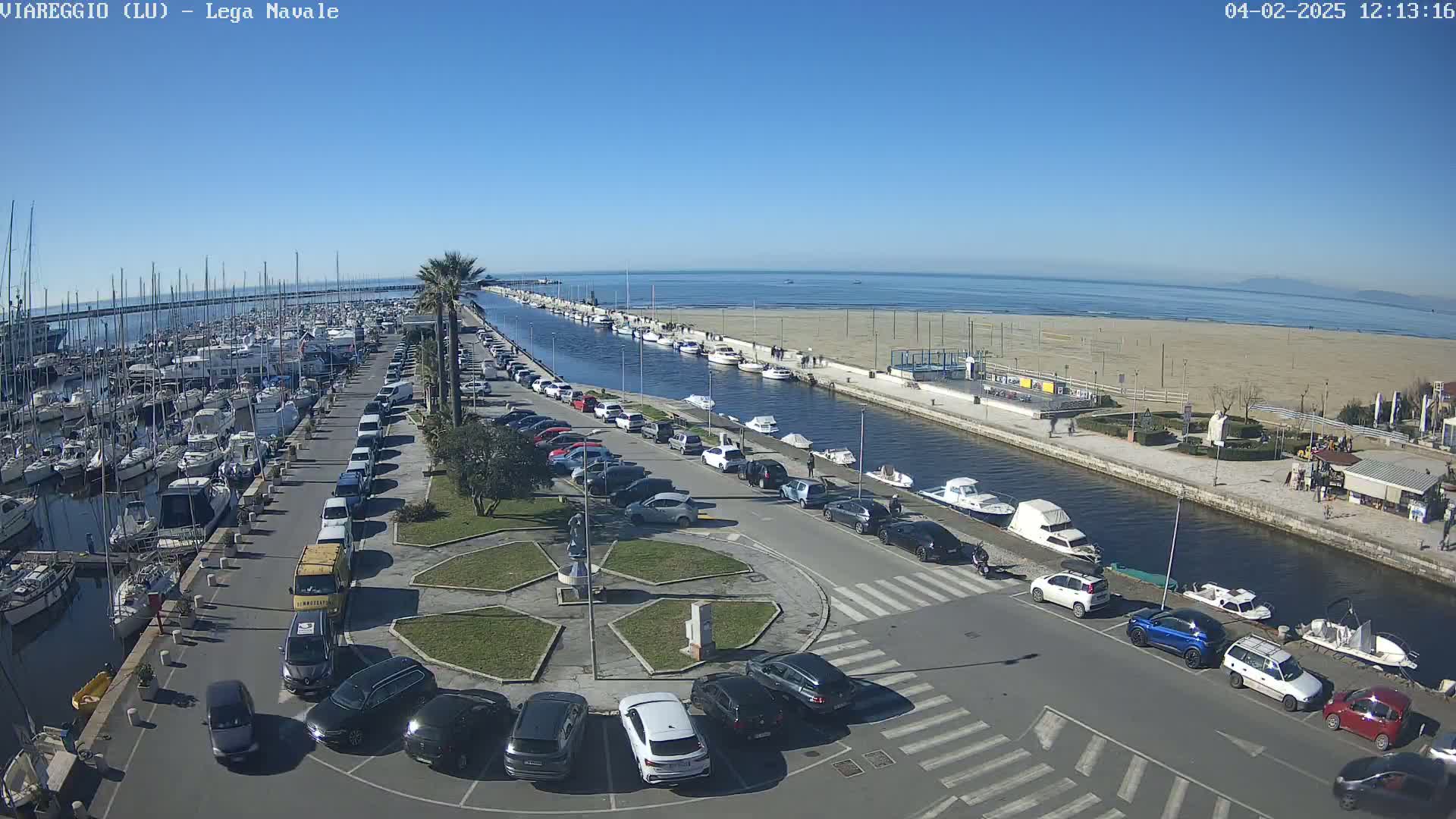 A marina filled with boats is next to a canal that leads to a sandy beach under a clear, sunny sky.