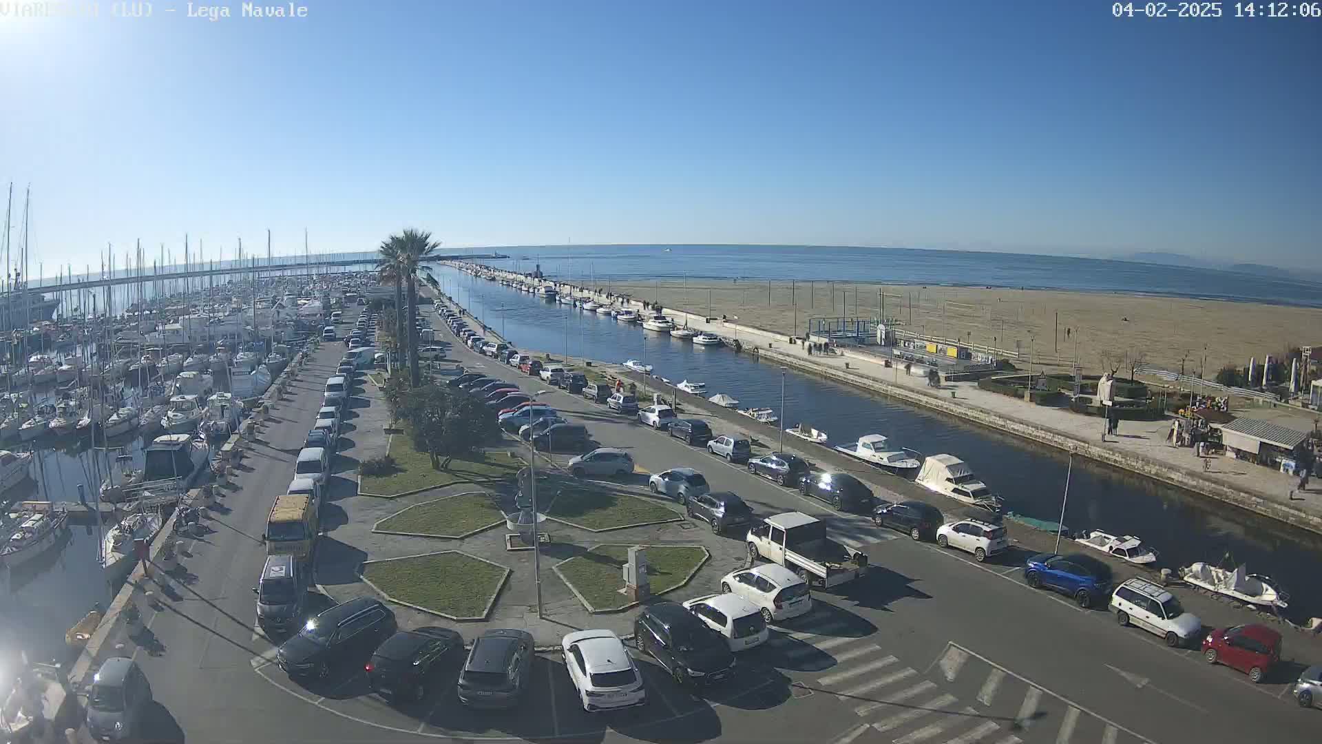 A marina filled with boats is next to a canal, with cars parked along the side and a sandy beach visible beyond the canal under a clear, sunny sky.