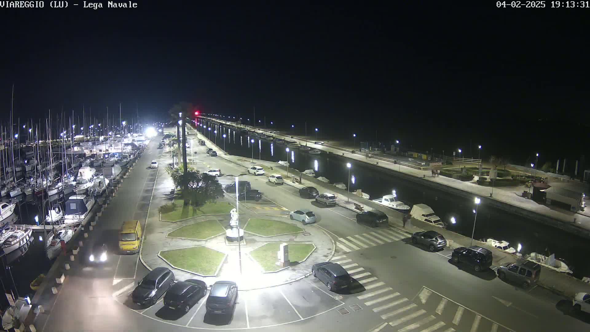 A nighttime aerial view of a marina with many boats docked, a parking lot with several cars, and a canal separating the marina from a paved walkway.