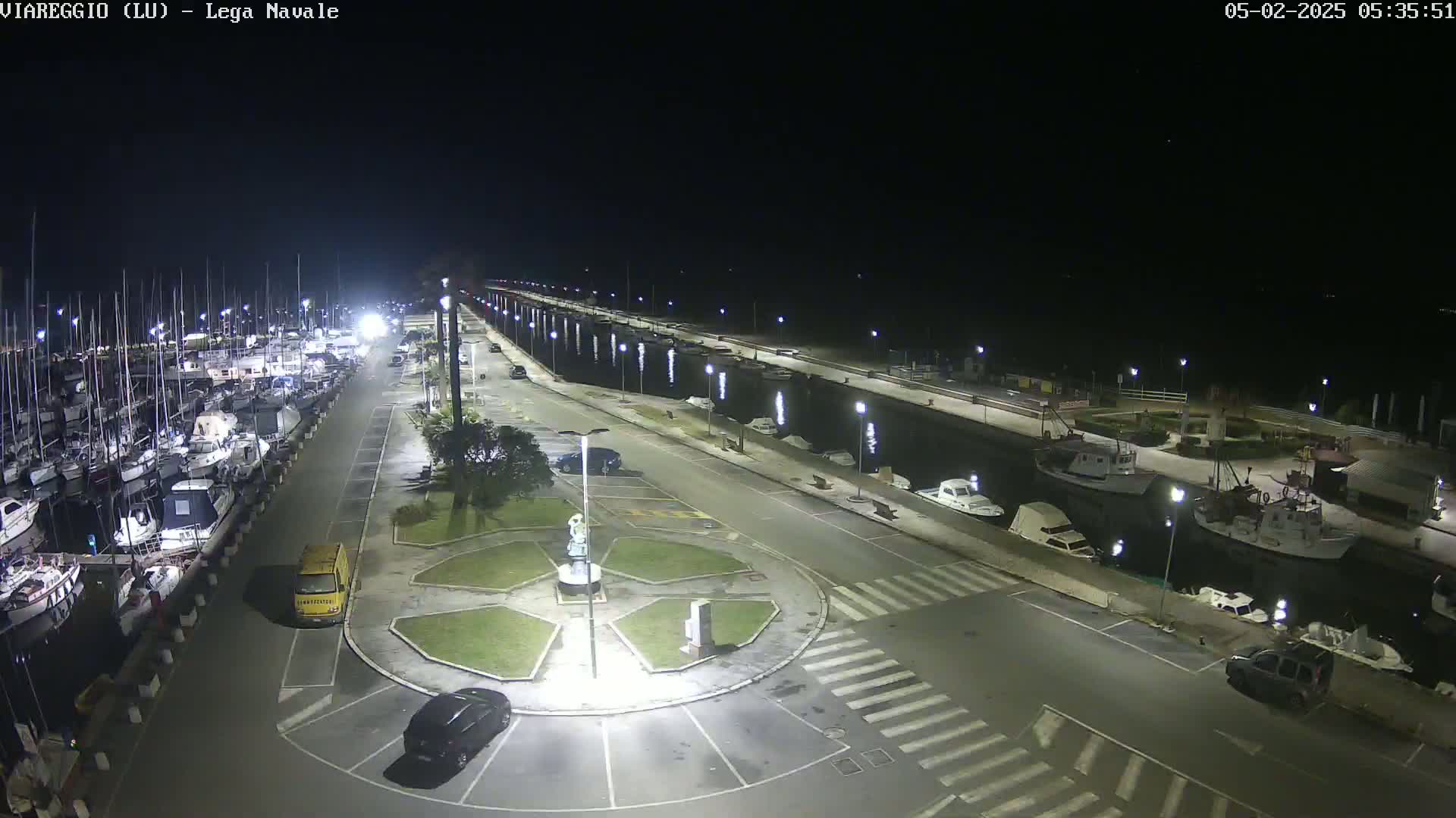 A nighttime aerial view of a marina with numerous boats docked, a paved area with cars parked, and a small roundabout in the center, illuminated by streetlights.