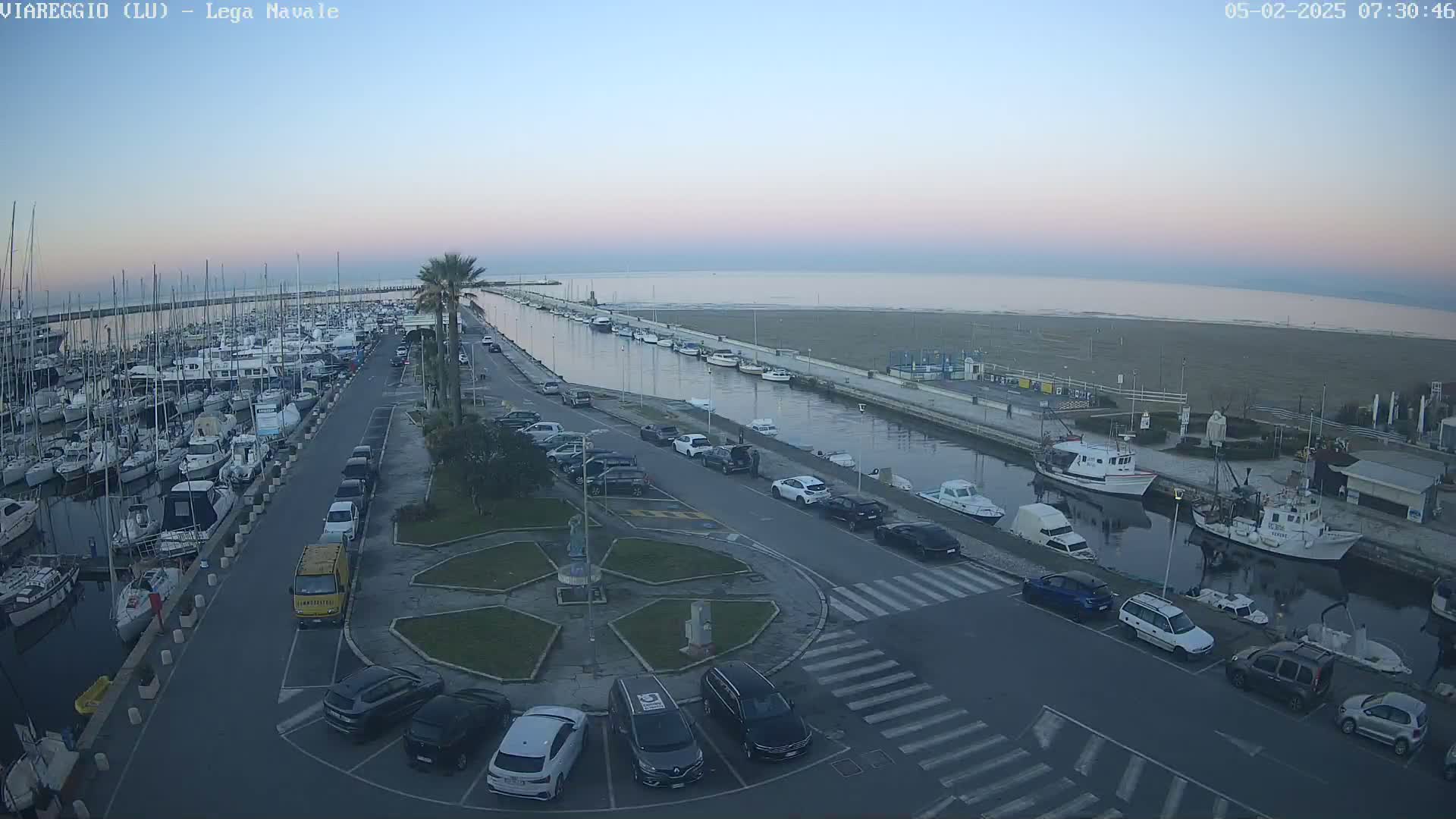 A marina filled with boats is next to a calm body of water under a twilight sky, with a parking lot and a roundabout nearby.