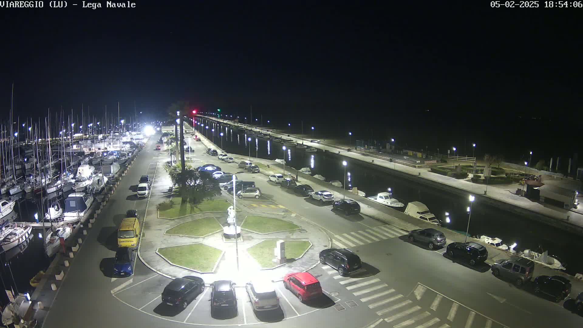A nighttime view of a marina with numerous boats docked, a parking lot filled with cars surrounding a landscaped roundabout, and a calm waterway extending into the distance under a clear, dark sky.
