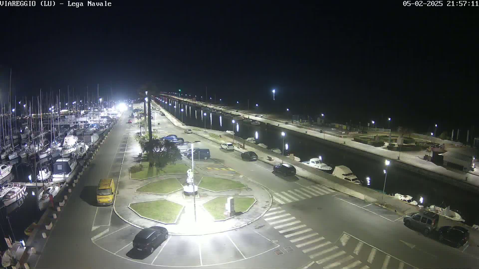 A nighttime view of a marina with numerous boats docked, a roadway with parked cars, and a central roundabout, all illuminated by streetlights.
