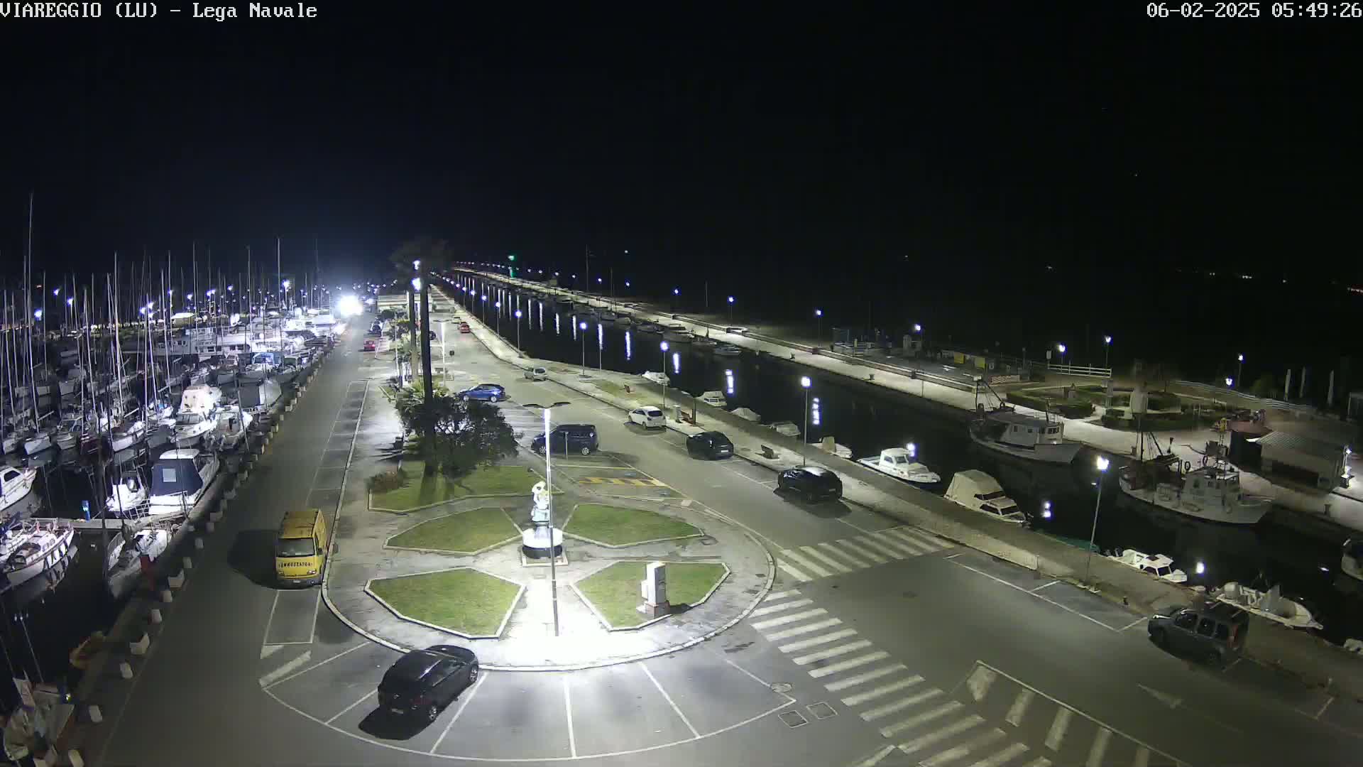 A nighttime aerial view of a marina with docked boats, cars parked in a lot, and a small roundabout in the foreground.