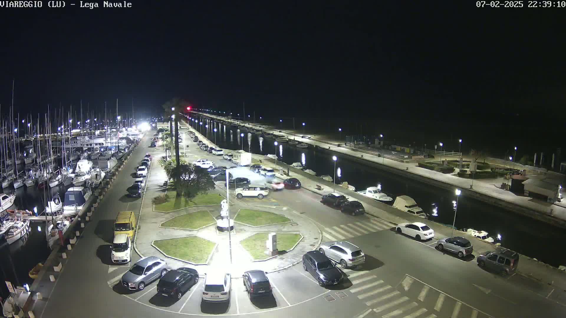 A nighttime aerial view shows a marina filled with boats, a parking lot with cars, and a canal with additional boats, all under a clear, dark sky.