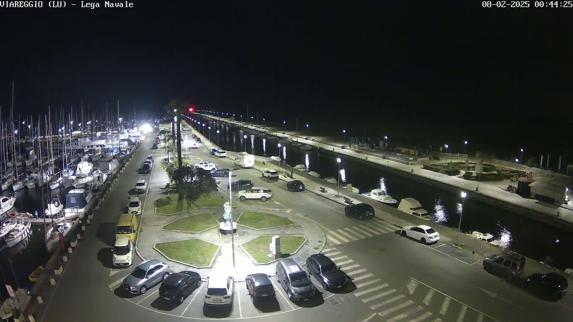 A nighttime aerial view of a marina with numerous boats docked, a parking lot filled with cars, and a calm canal reflecting lights.