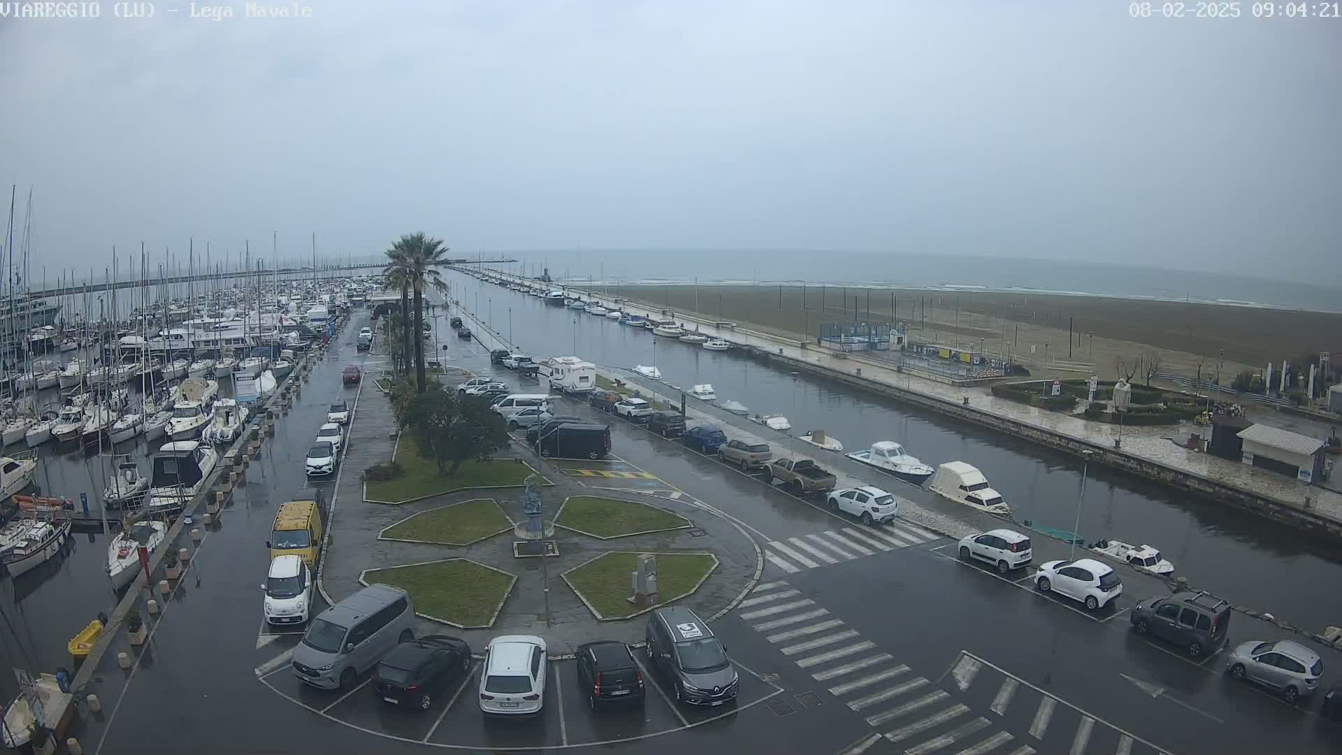A wet harbor filled with numerous boats is adjacent to a parking lot and a canal leading to a beach under an overcast sky.