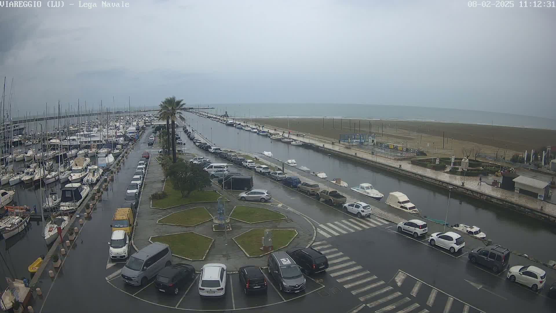 A marina filled with boats is shown on a cloudy day, with a canal separating it from a wet parking lot and beach.
