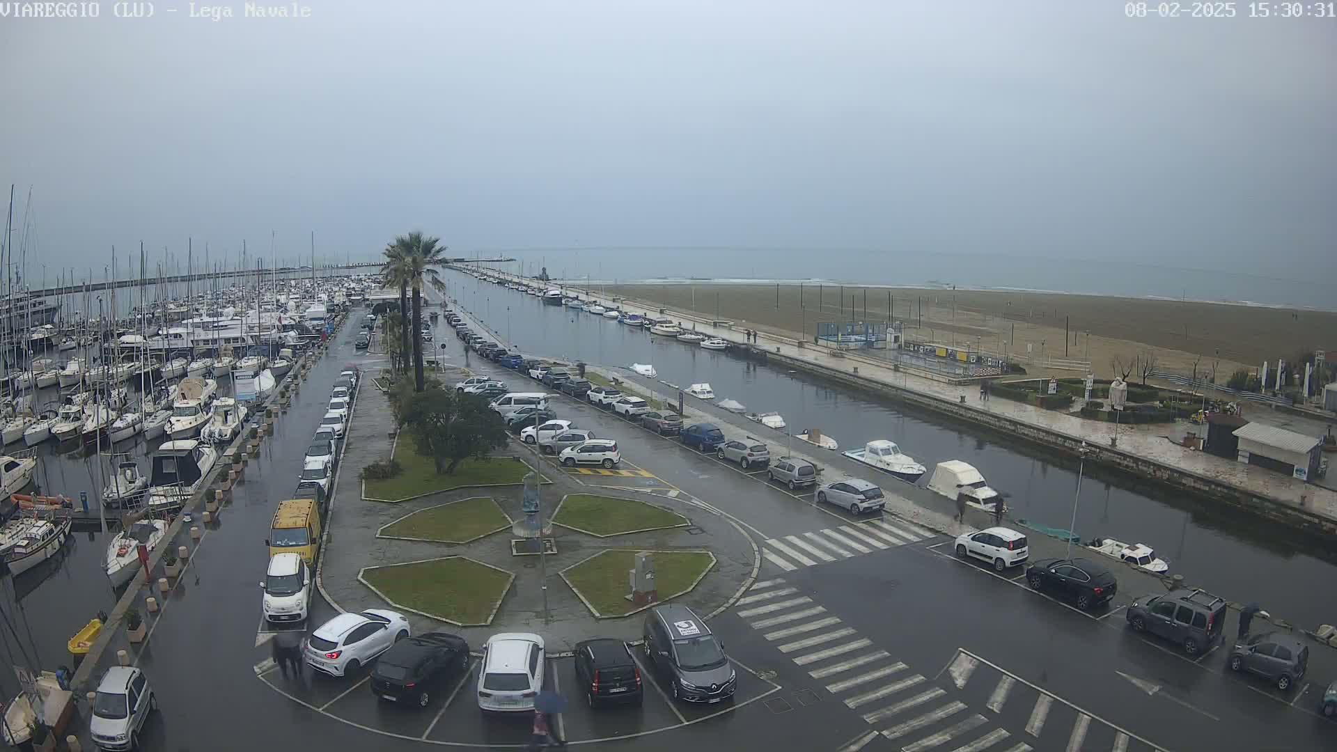 A marina filled with boats is shown next to a canal and sandy beach on an overcast day, with cars parked along the canal's edge.