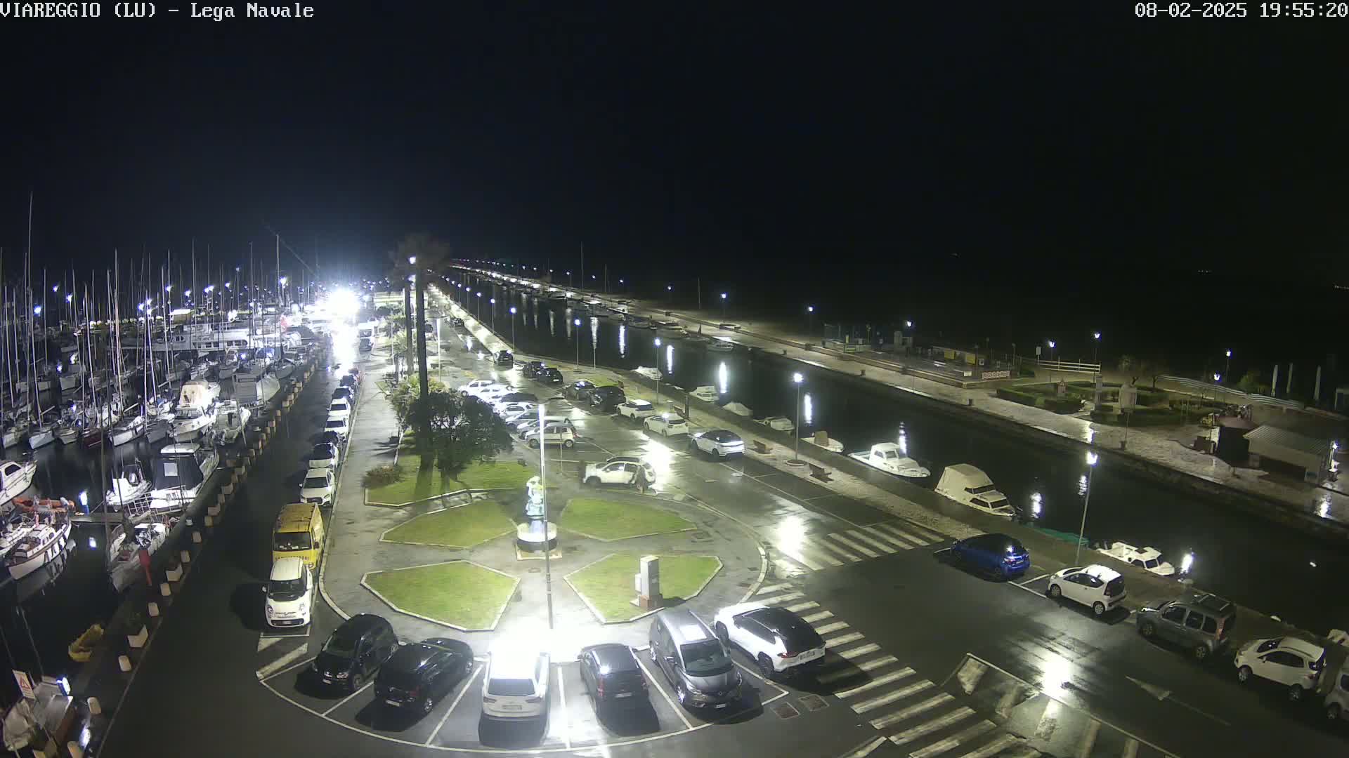 A nighttime, wet marina scene shows numerous boats docked along a canal, with cars parked in a lot adjacent to a landscaped area.