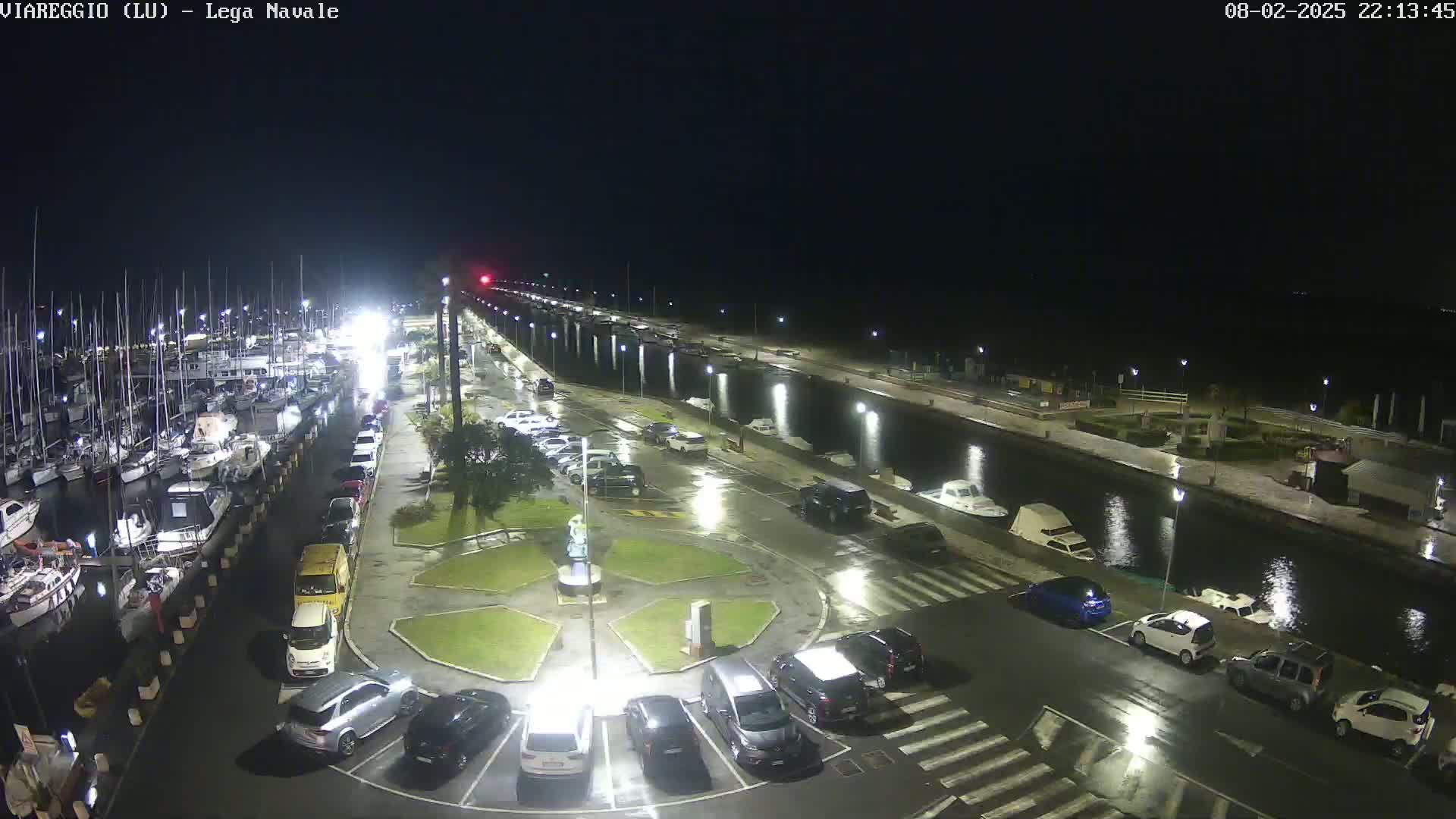 A nighttime view of a marina with many boats docked, a wet parking lot filled with cars, and a canal reflecting lights.