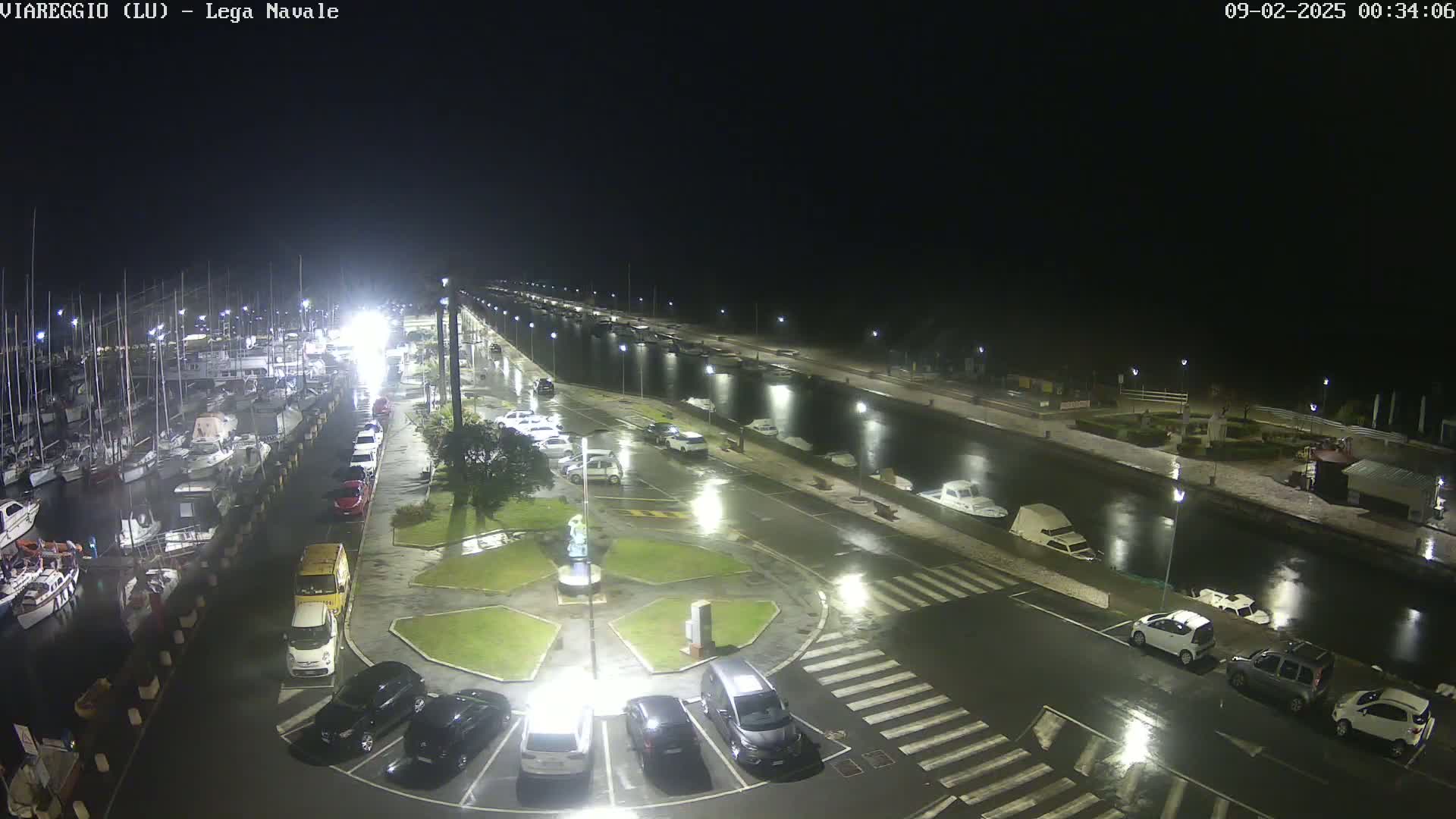 A nighttime view of a marina with numerous boats docked, a wet parking lot filled with cars, and a canal reflecting lights under a rainy sky.
