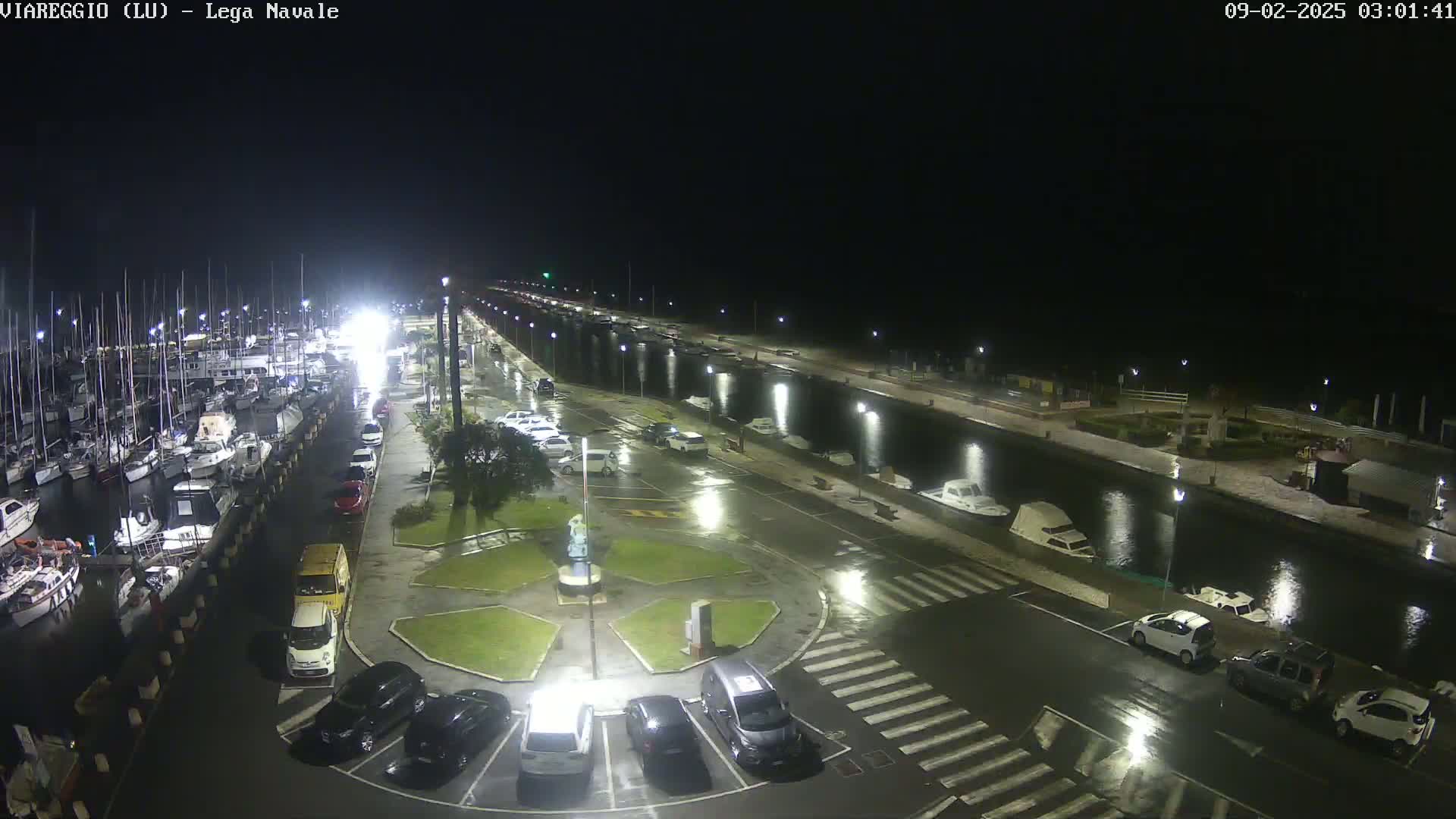 A nighttime, rain-slicked marina and parking lot scene shows numerous boats docked, cars parked, and water reflecting lights.