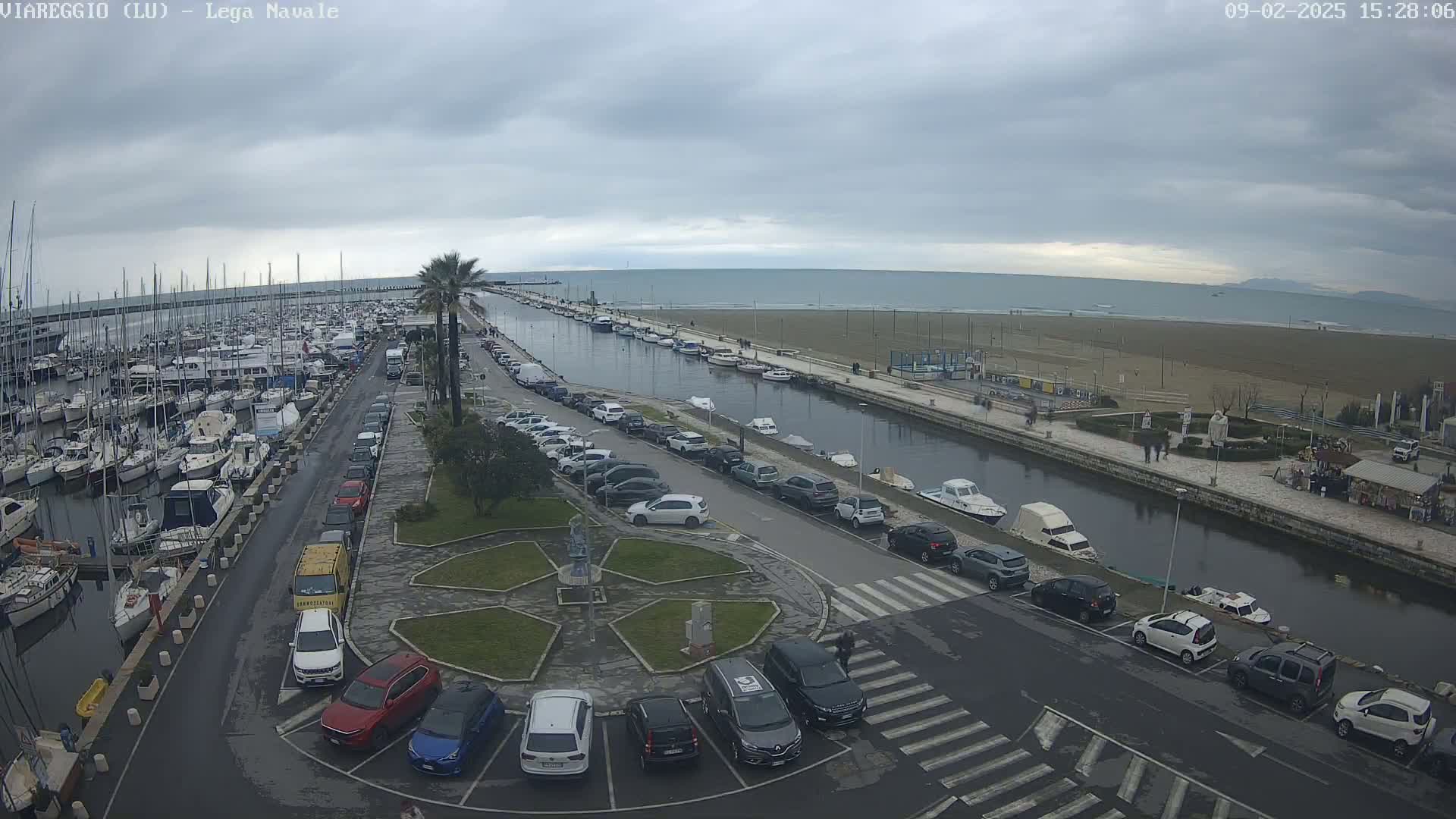 A marina filled with boats is next to a canal and a beach under an overcast sky.