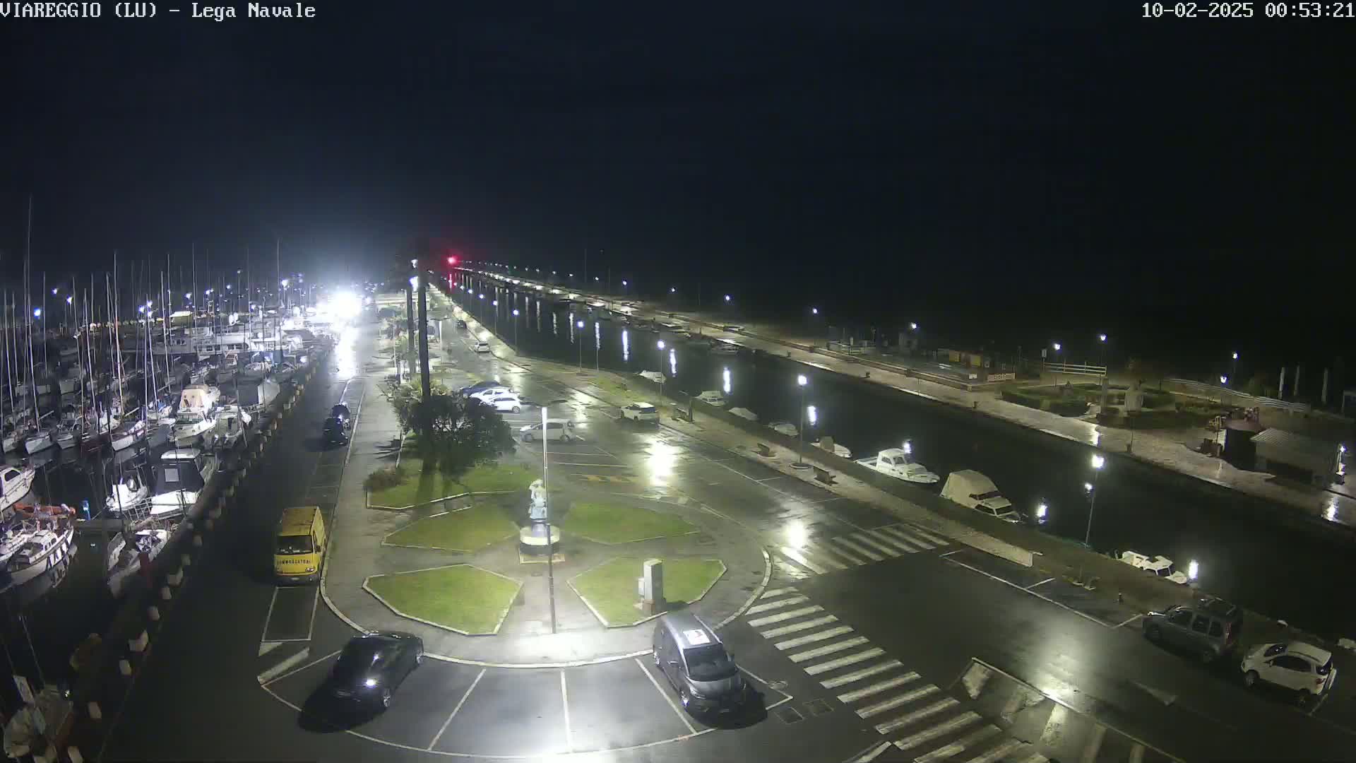 A nighttime view of a marina with docked boats, wet streets, and several cars parked in a lot under lightly rainy conditions.