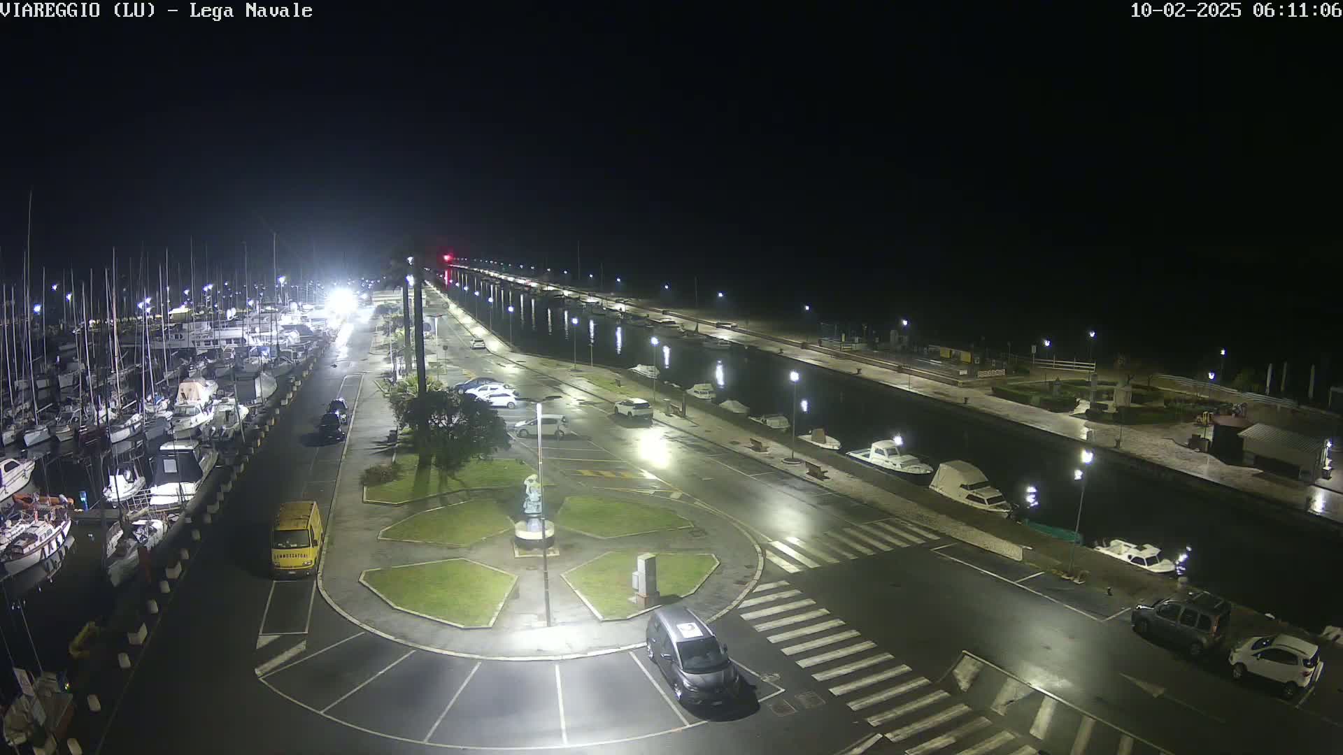 A nighttime, wet marina scene shows numerous boats docked along a paved walkway lined with streetlights, with cars parked in a nearby lot and a roundabout in the foreground.