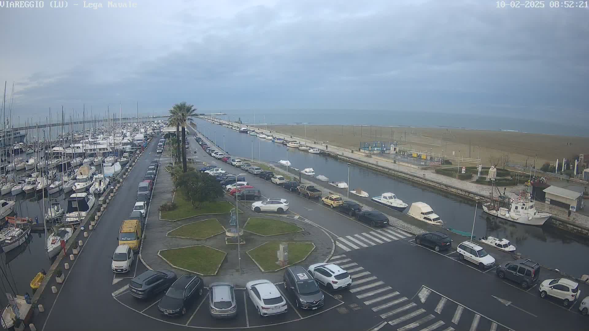 A marina filled with boats is next to a canal and beach under an overcast sky.