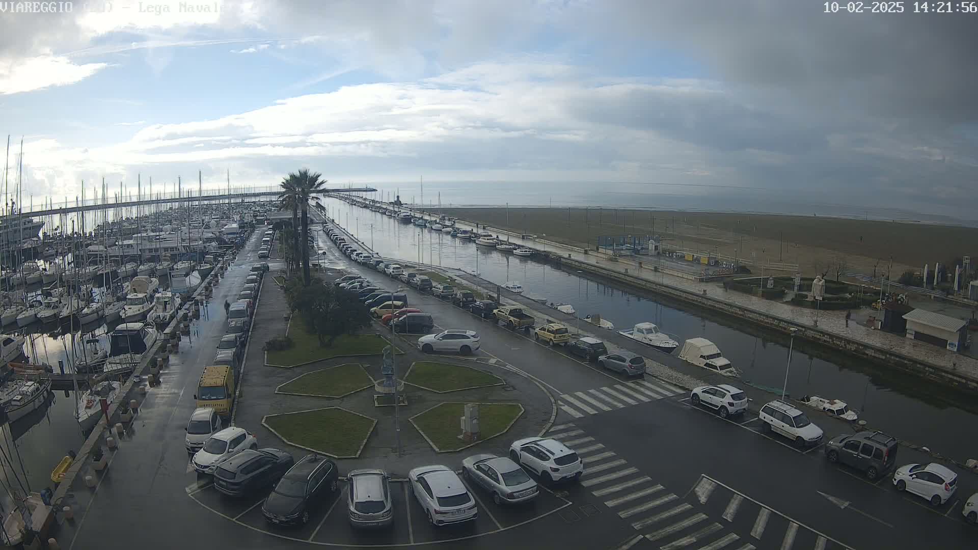 A wet marina with many boats and cars parked in a lot, next to a calm waterway leading to a sandy beach under a mostly cloudy sky.