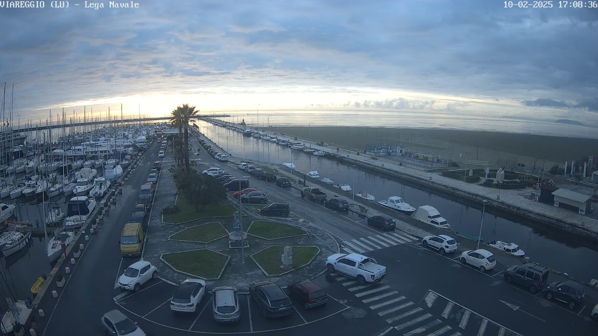 A marina filled with boats is next to a canal, which leads to a beach under a partly cloudy sky.