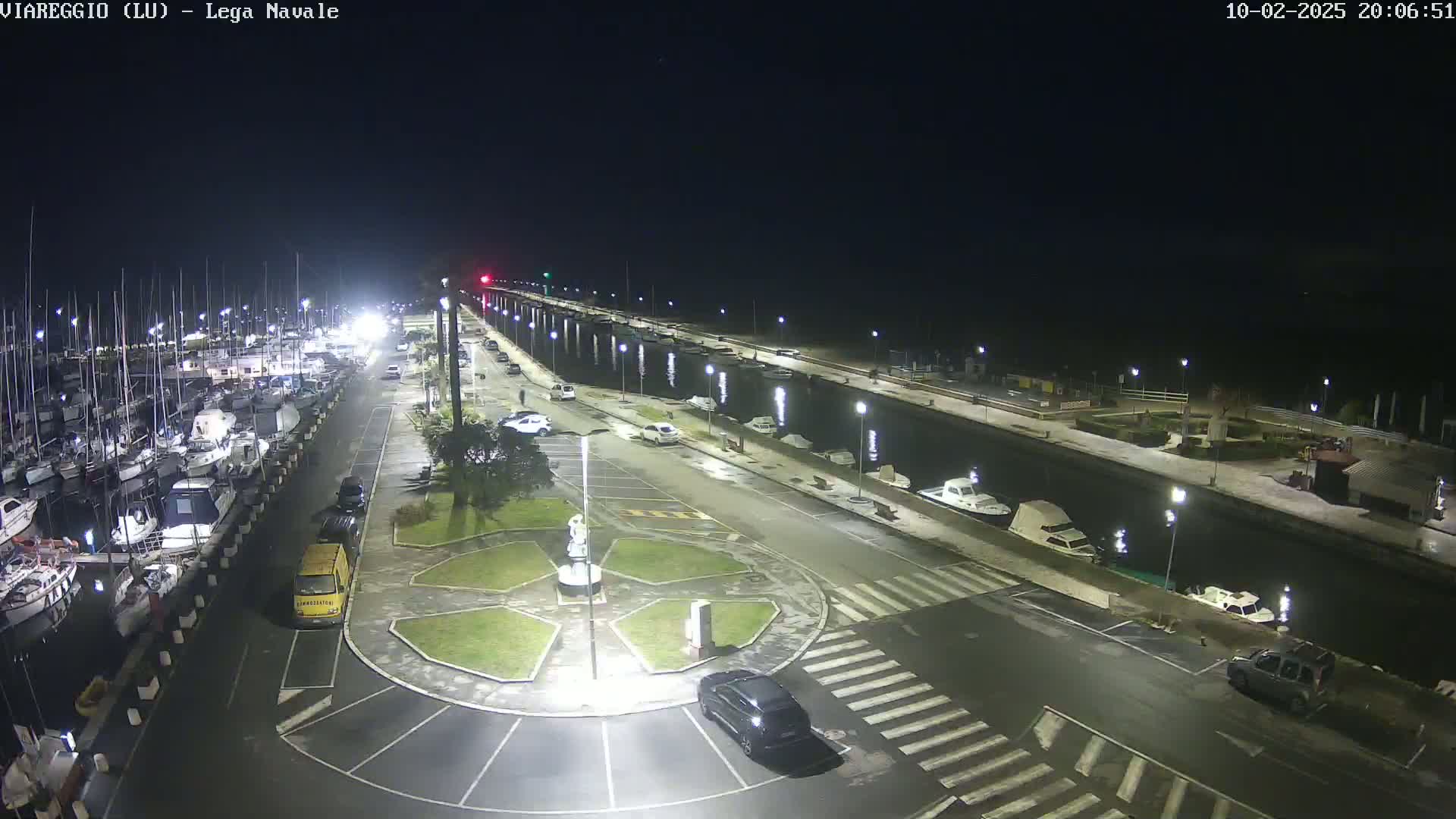 A nighttime aerial view of a marina with numerous boats docked, cars parked in a lot, and a canal running alongside a roadway.