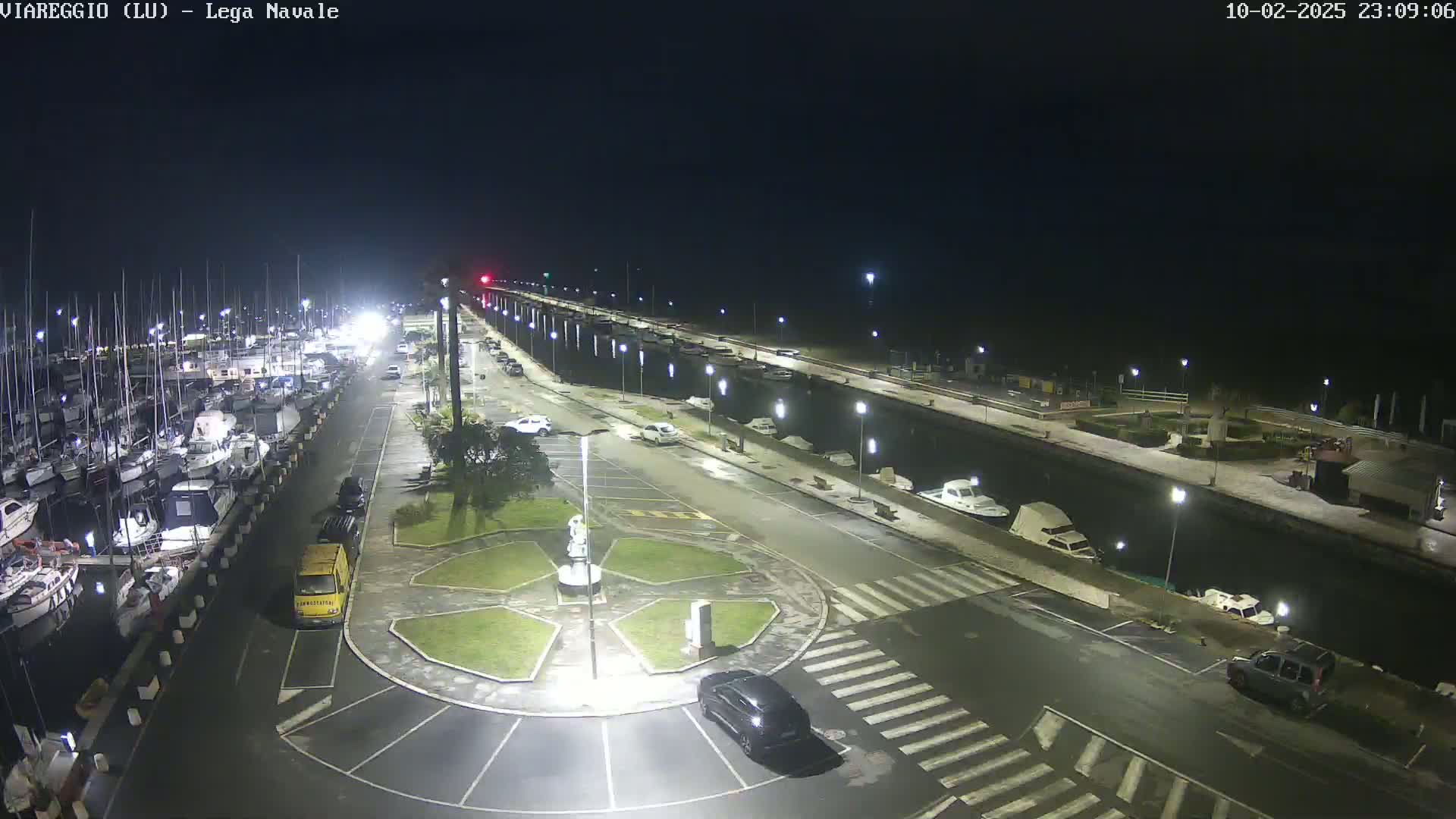 A nighttime, illuminated view of a marina with numerous boats docked alongside a roadway, featuring a small roundabout and several parked cars.