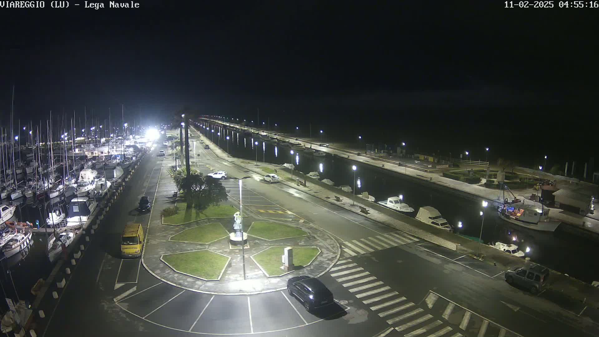 A nighttime, well-lit marina with numerous boats docked along a paved waterfront, featuring a small roundabout and several cars parked nearby.