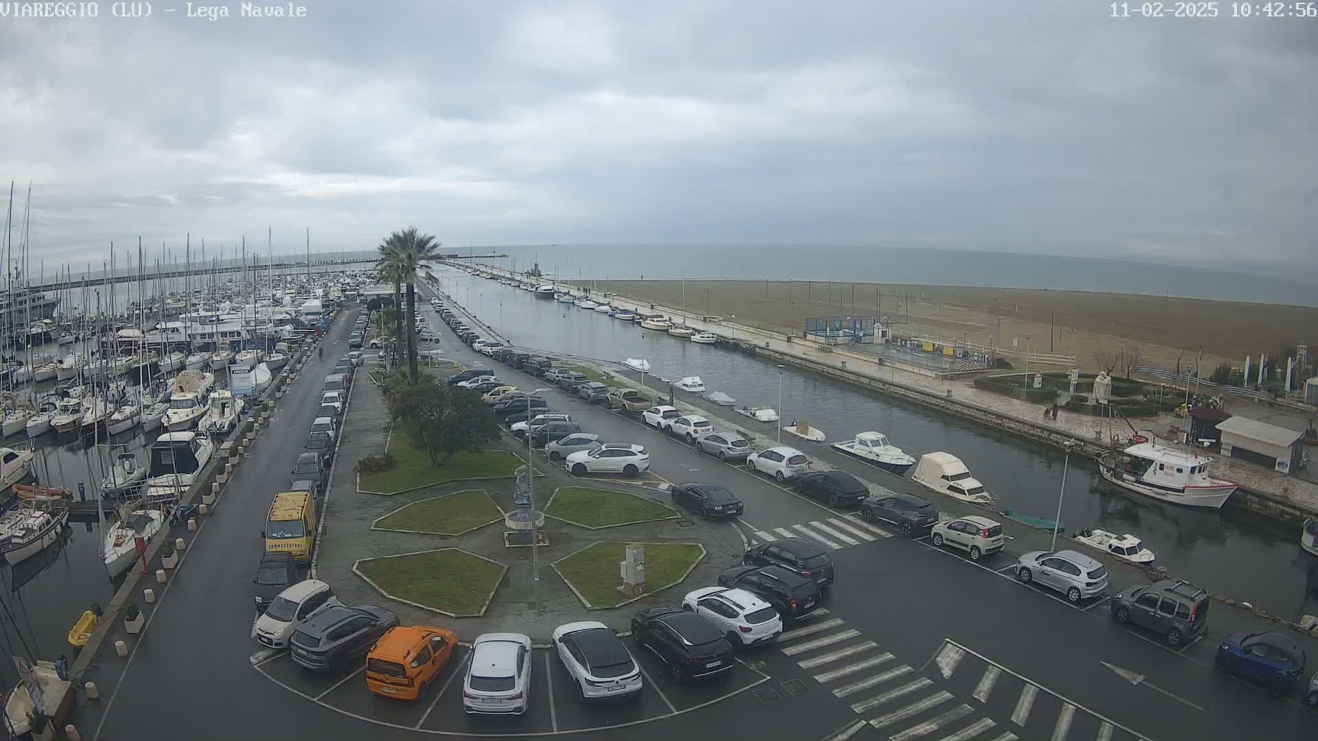A harbor filled with boats and yachts is next to a sandy beach under an overcast sky, with a canal separating the harbor and beach, lined with parked cars.