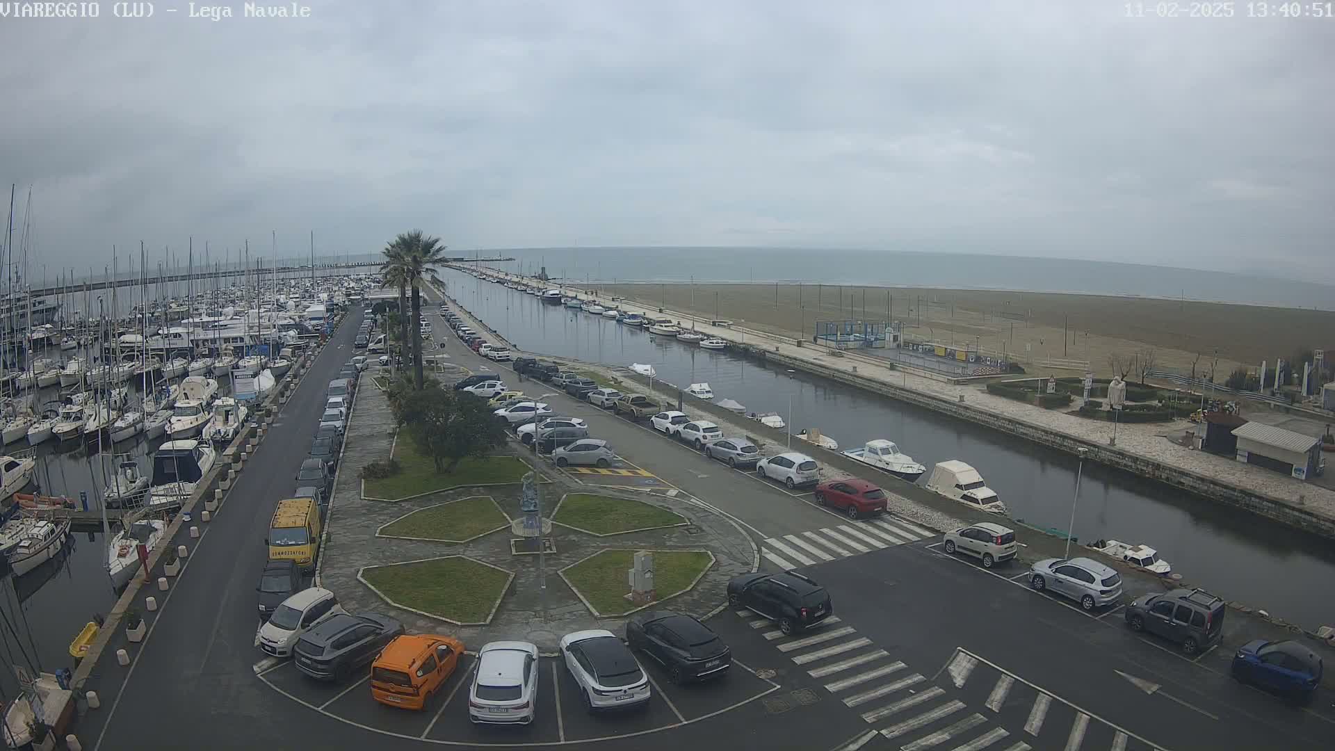 A marina filled with boats is next to a canal lined with parked cars, all situated beside a beach under an overcast sky.