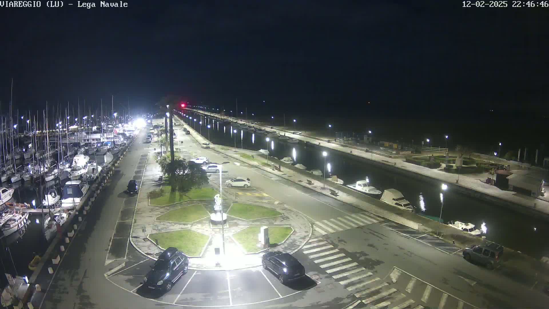 A nighttime aerial view of a marina with numerous boats docked, cars parked in a lot nearby, and a canal running alongside a paved walkway.