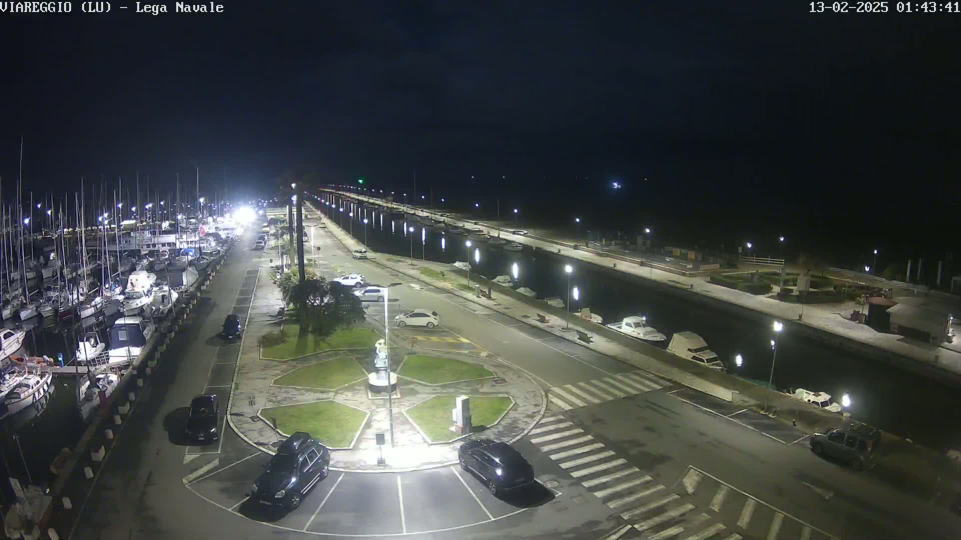 A nighttime, overhead view of a marina with docked boats, cars parked in a lot, and a canal under clear skies.