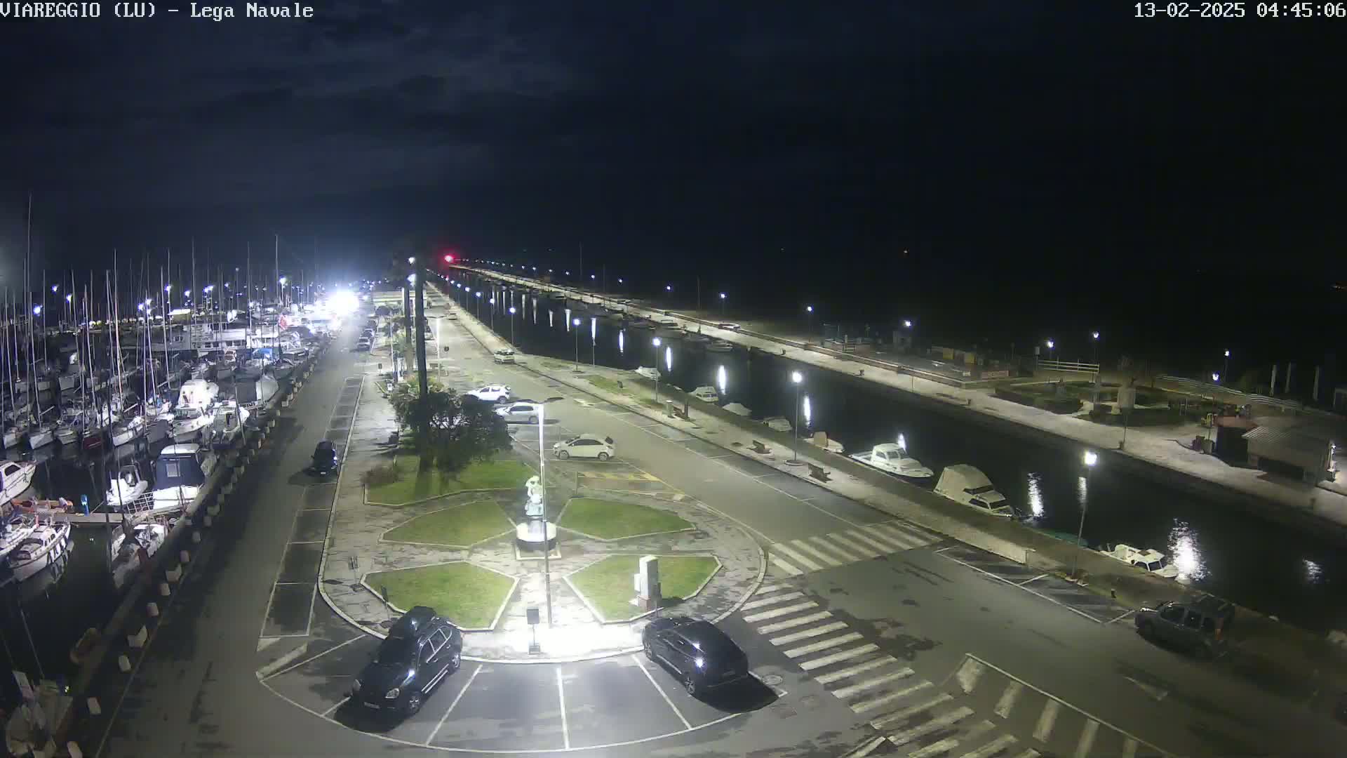 A nighttime view of a marina with numerous boats docked, cars parked in a lot nearby, and a calm canal reflecting lights.