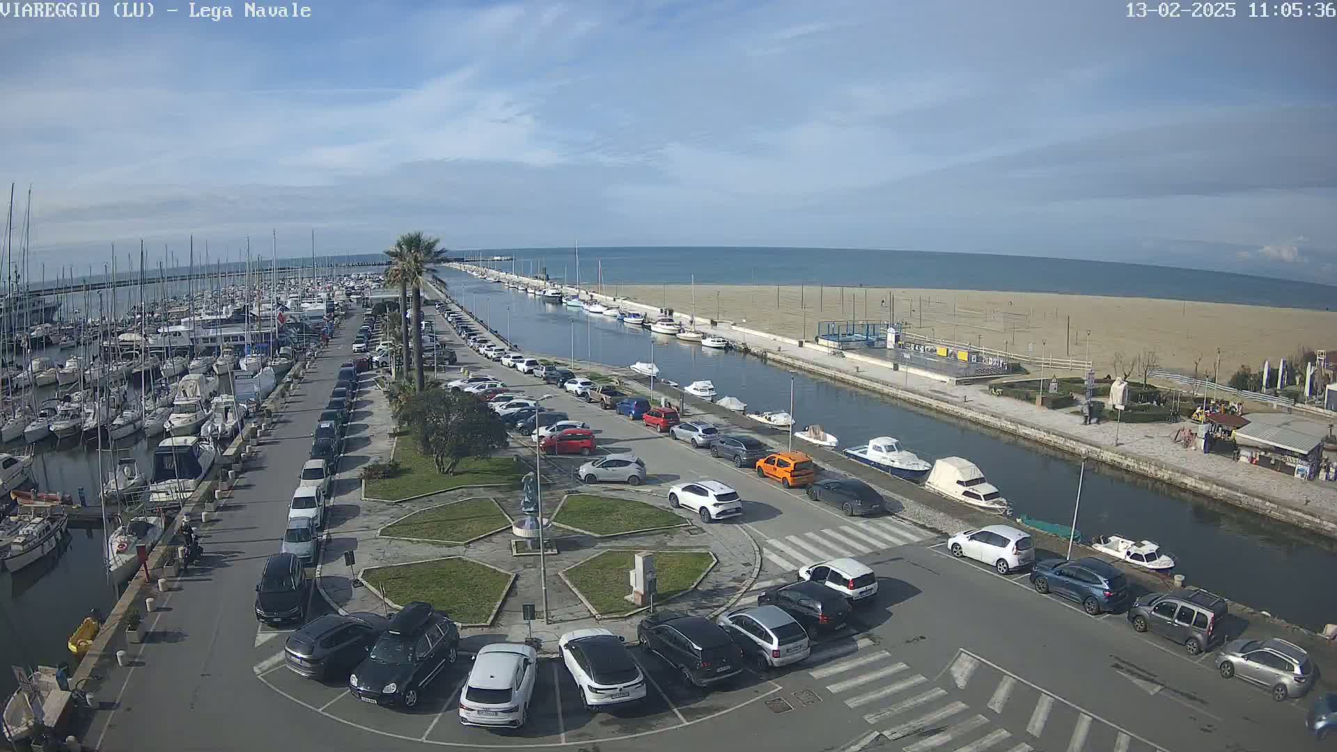 An aerial view shows a marina filled with boats, a canal, a sandy beach, and a parking lot full of cars under a mostly clear sky.