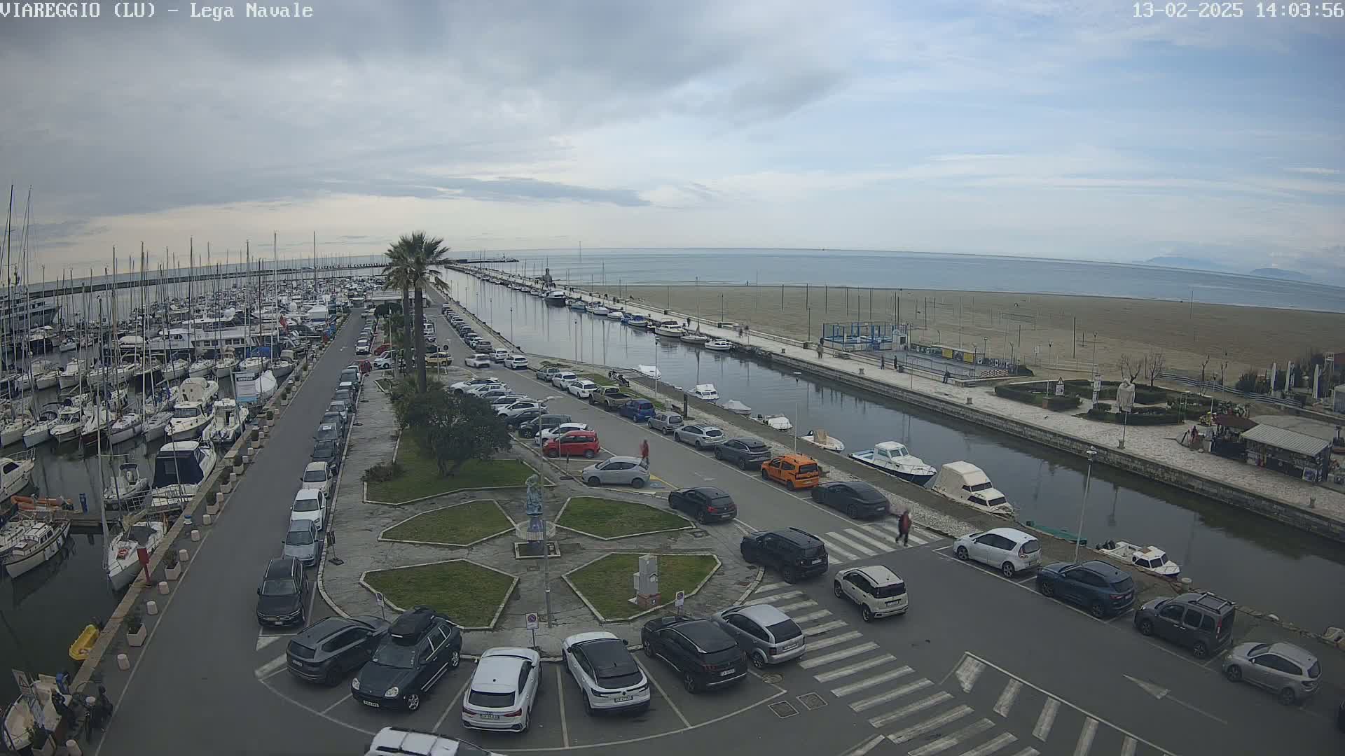 An aerial view shows a marina filled with boats, a canal, a sandy beach, and a parking lot full of cars, all under a mostly cloudy sky.